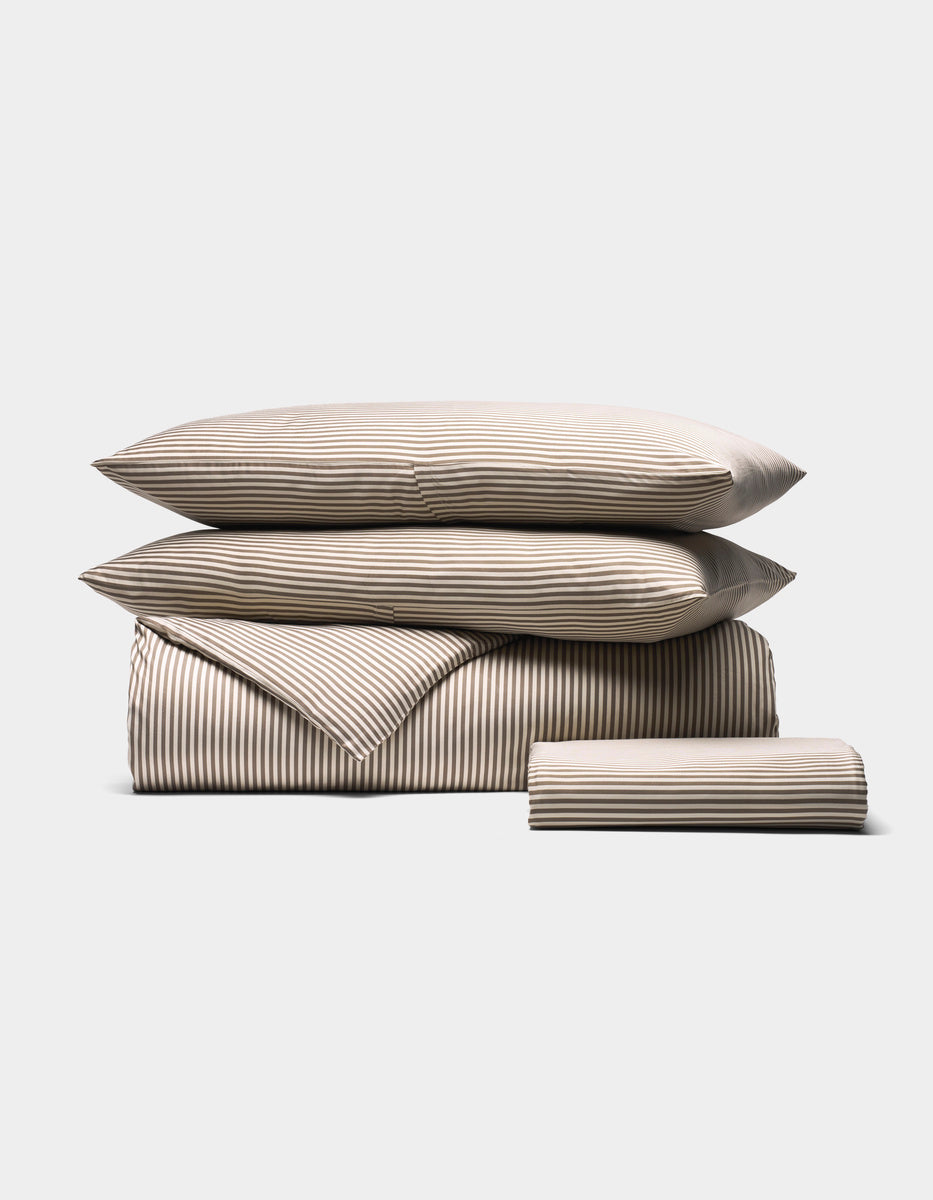 The Cozy Earth Bamboo Bedding Essential Bundle features beige and white striped bedding—two pillows, a folded duvet, and a folded sheet—neatly displayed against a plain light gray background.