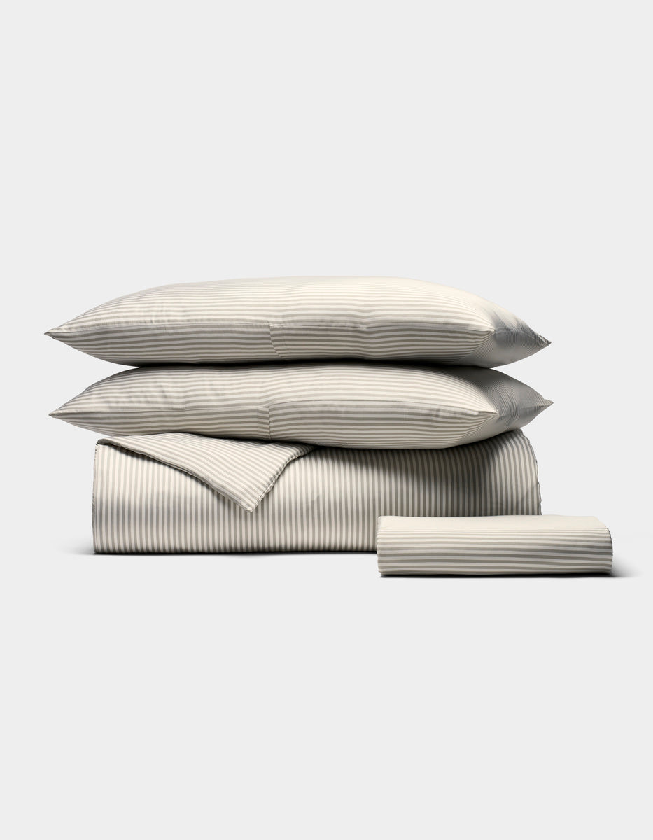 The Bamboo Bedding Essential Bundle by Cozy Earth features a neatly stacked set of beige and white striped bedding—two pillows, a folded duvet, and a folded sheet—displayed on a plain light background.