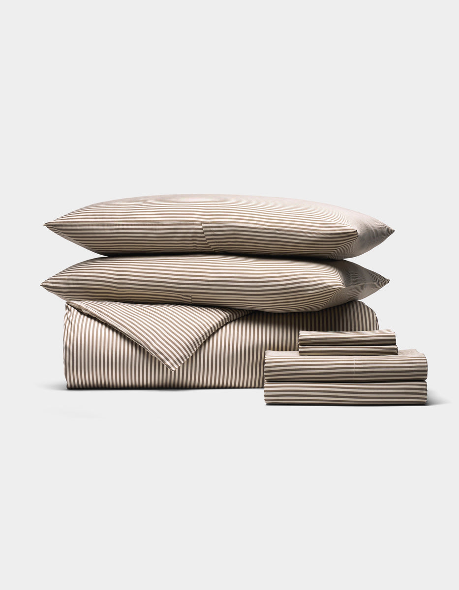 The Cozy Earth Bamboo Bedding Ultimate Bundle features two pillows, a comforter, and sheets in beige and white horizontal stripes, all neatly folded and arranged on a white background.