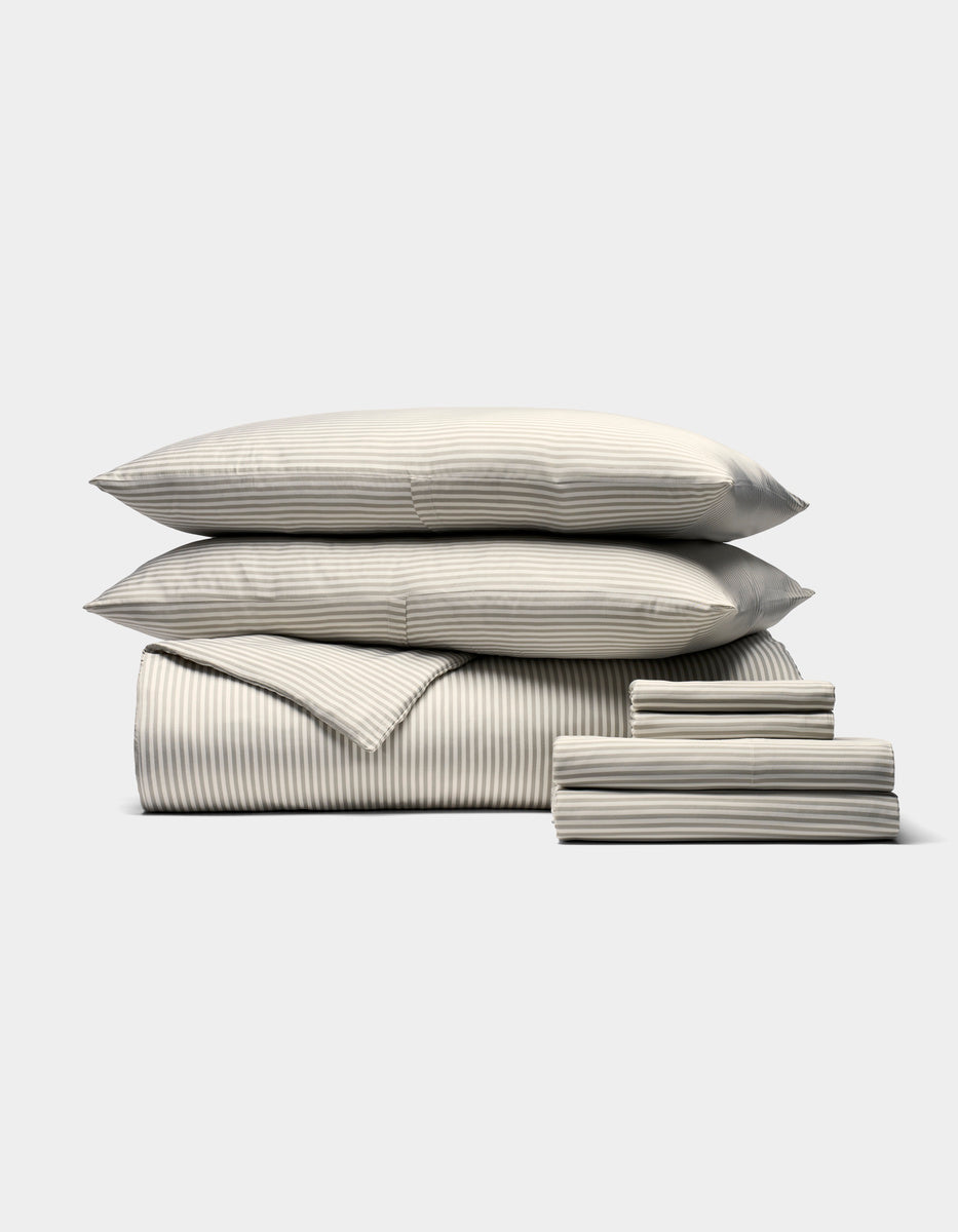 The Cozy Earth Bamboo Bedding Ultimate Bundle includes pillows, a duvet, and sheets in a light cream and grey pinstripe design, neatly stacked and displayed against a plain white background.