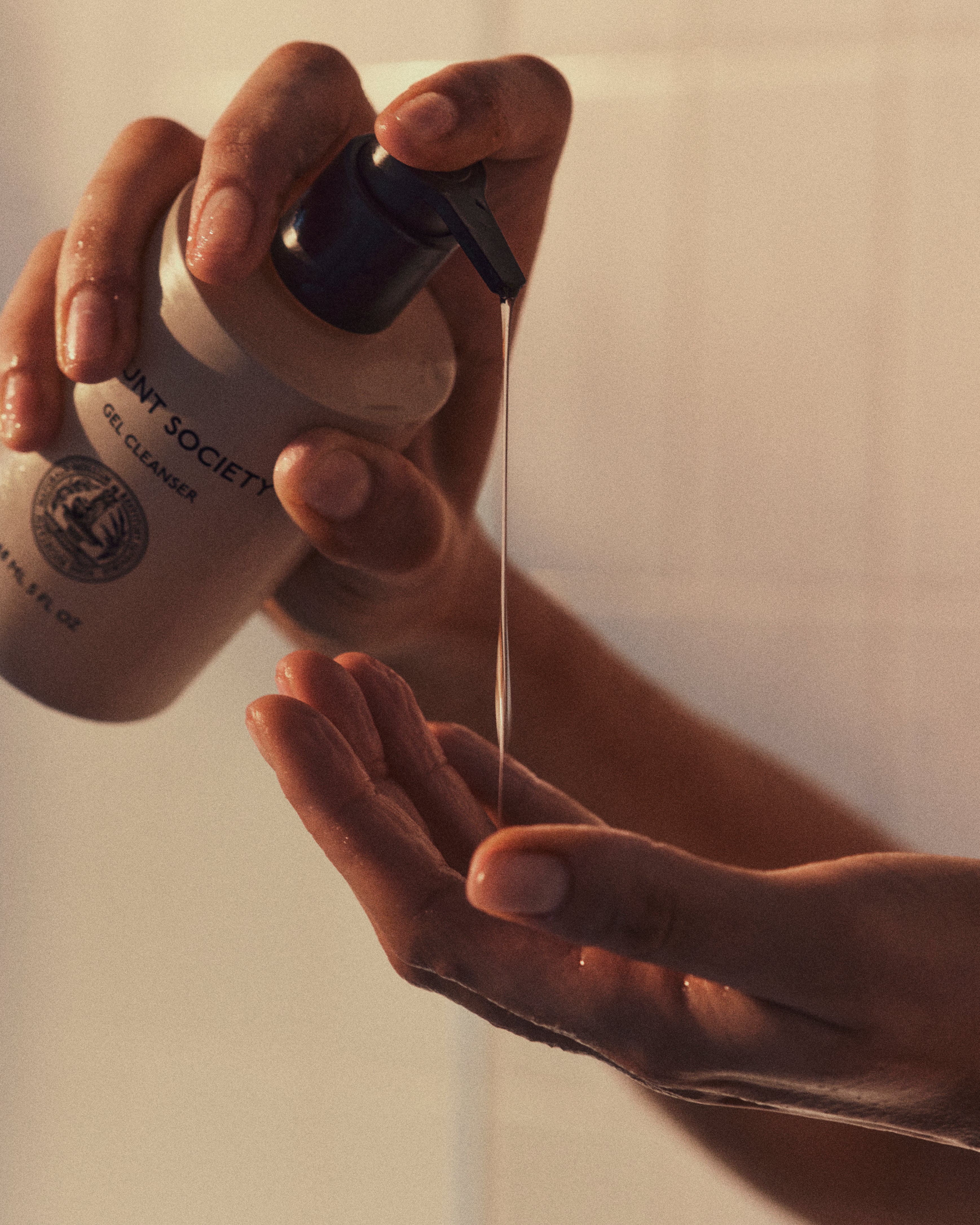Close-up of hands dispensing Cozy Earth's Gel Cleanser from a pump bottle. One hand is holding the bottle and pressing the pump, while the other hand is positioned below to catch the gel. The background is softly lit with warm tones.
