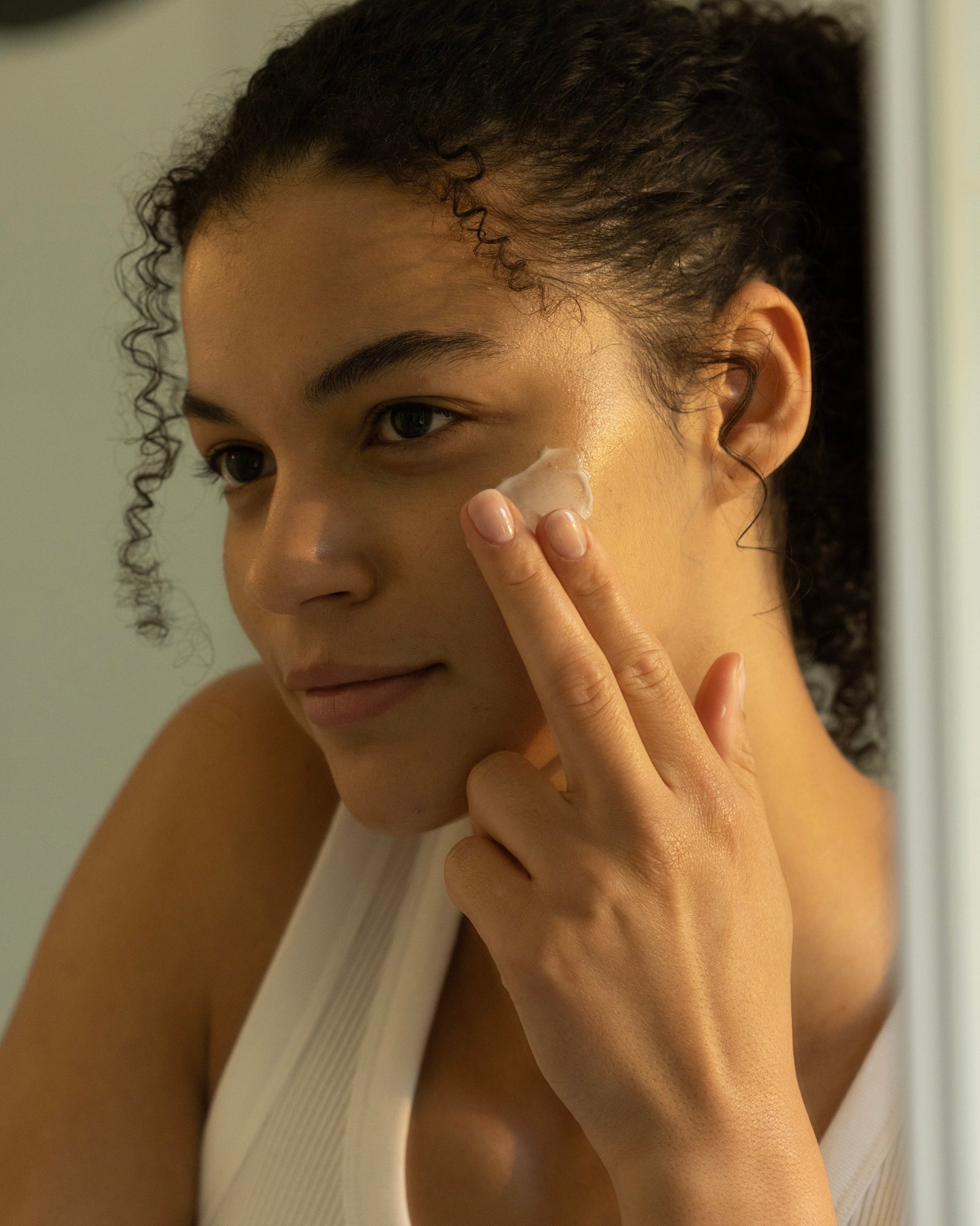 A curly-haired individual applies a dollop of The Hydration Duo by Cozy Earth to their cheek using their fingers. They are looking into a mirror, dressed in a sleeveless white top, and appear deeply focused on their skincare routine. The background is softly lit.