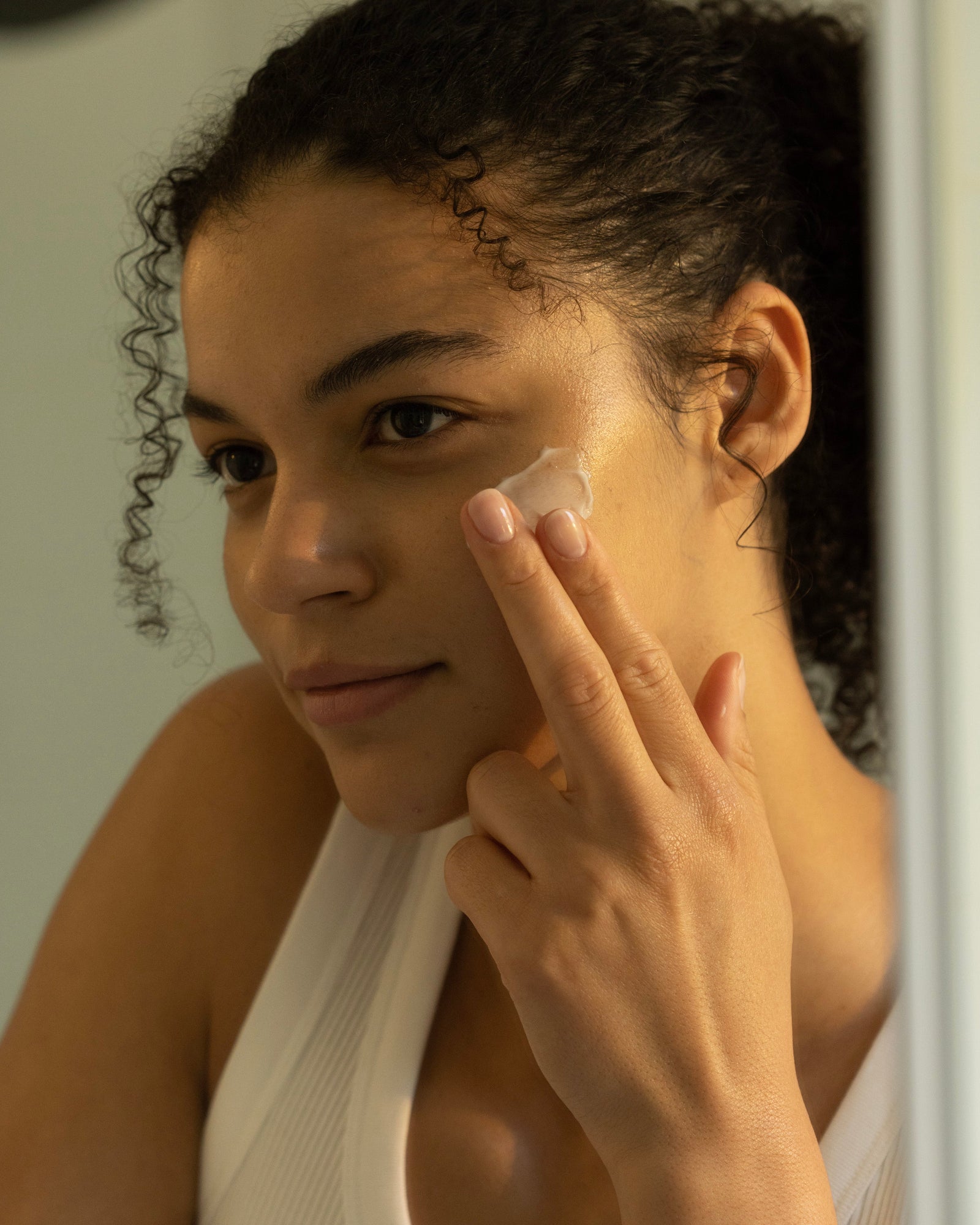 A person with curly hair, wearing a white tank top, dabs Cozy Earth's "Water Cream Moisturizer" cream on their cheek with their fingertips, seemingly engaged in a skincare routine.