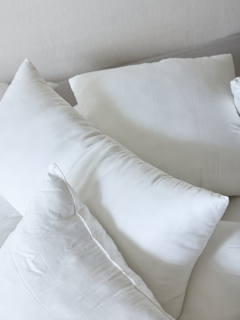 A close-up view of several **Cozy Earth Premium Down Alternative Inserts** arranged on a bed with white sheets. The pillows, varying slightly in shape and size, create a soft and cozy atmosphere. The background features a muted light color, enhancing the serene and clean setting.