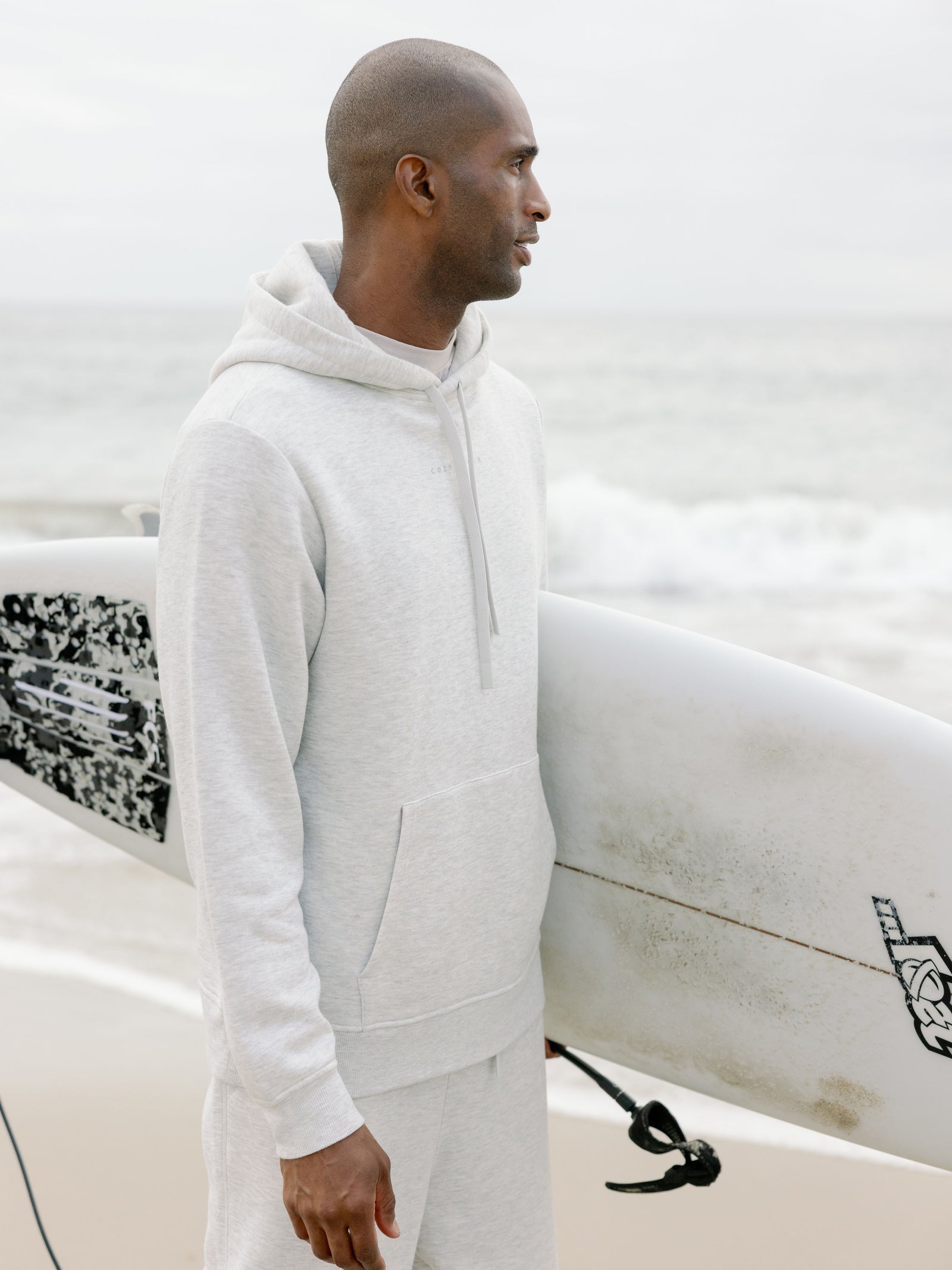 Man wearing heather grey cityscape hoodie holding surfboard 
