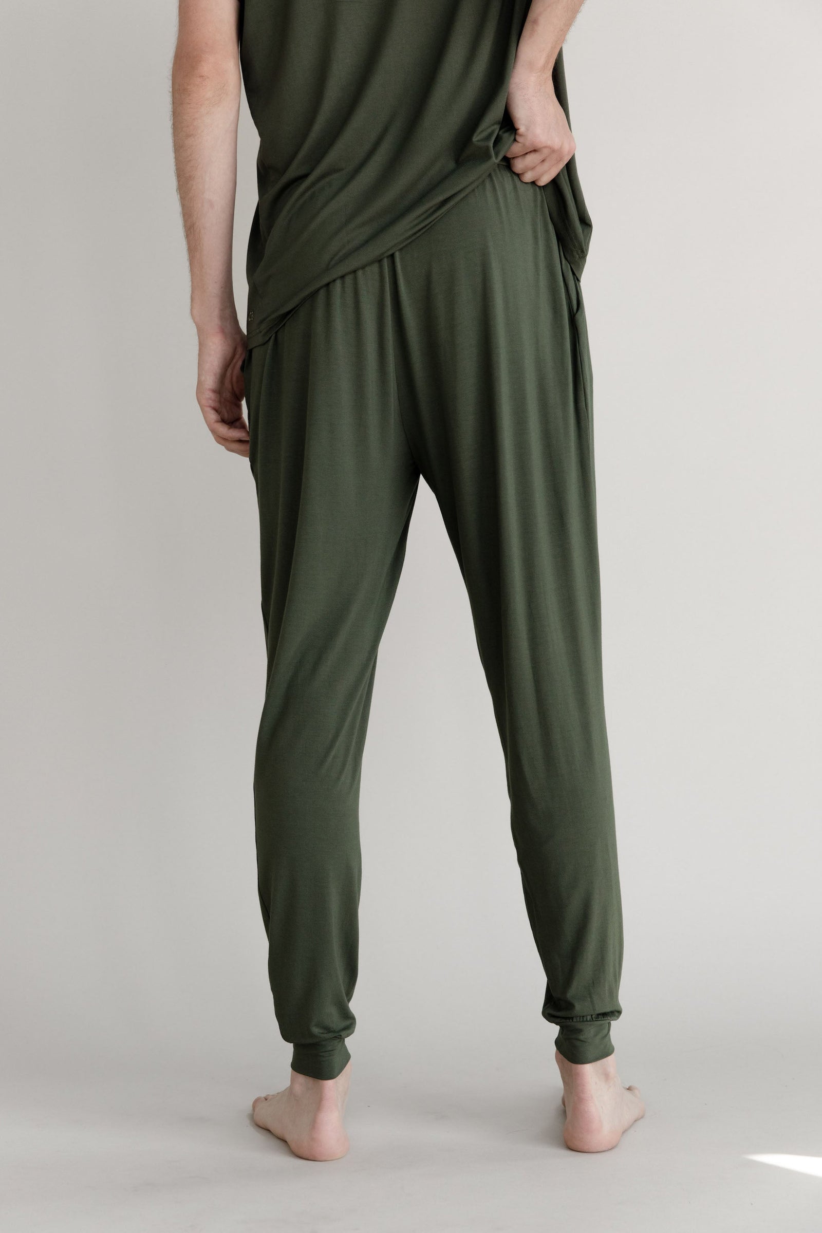 A person wearing the Men's Stretch-Knit Long Sleeve Bamboo Pajama Jogger Set from Cozy Earth in olive green is standing barefoot against a plain light gray background. The person is slightly turned away from the camera, with one hand reaching behind their back. 