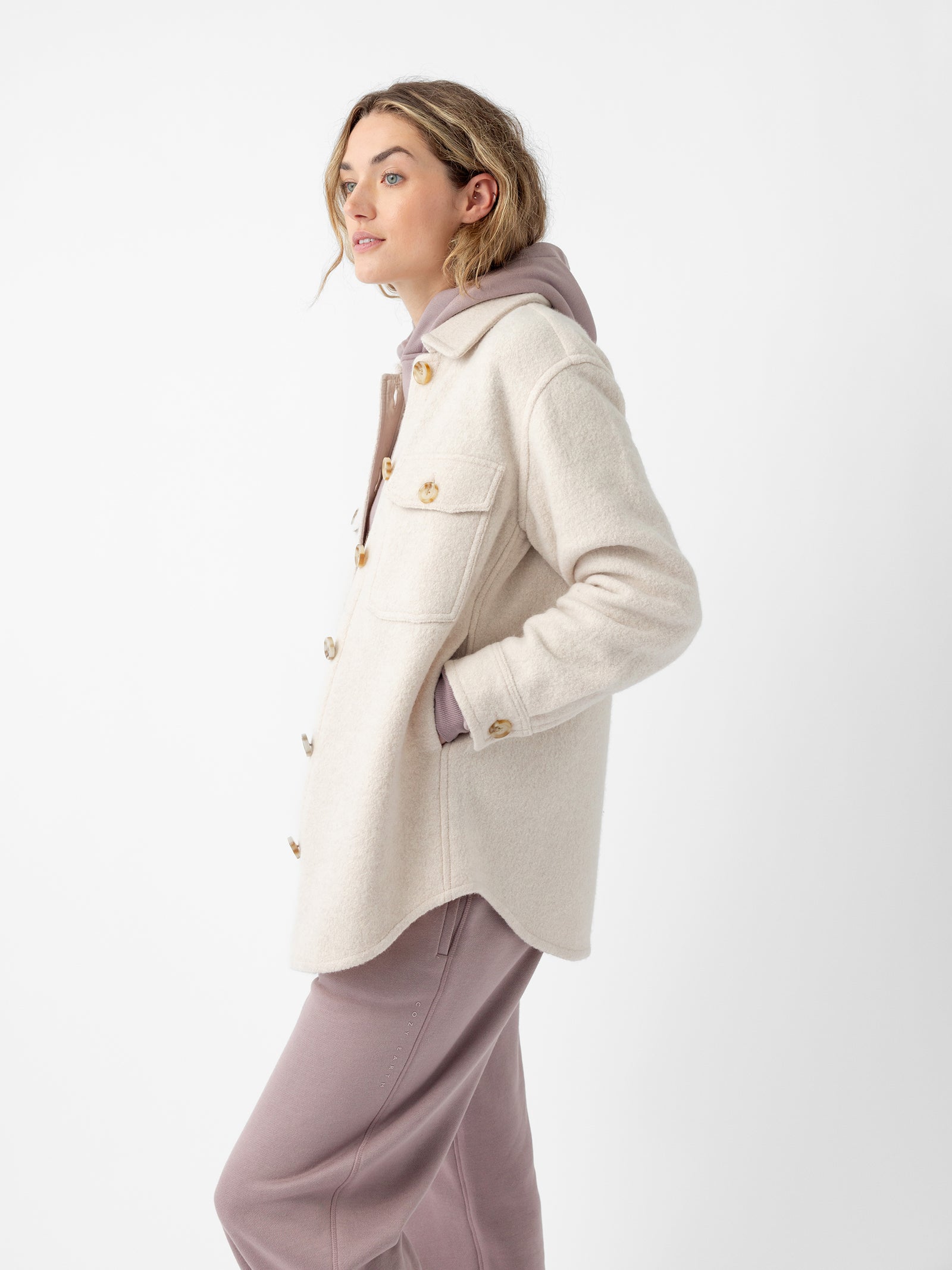 A person with shoulder-length light hair stands against a plain white background. They are wearing Cozy Earth’s Women’s Boucle Shacket in cream over a mauve hoodie, paired with matching mauve pants. The person looks to the side with a relaxed expression.  