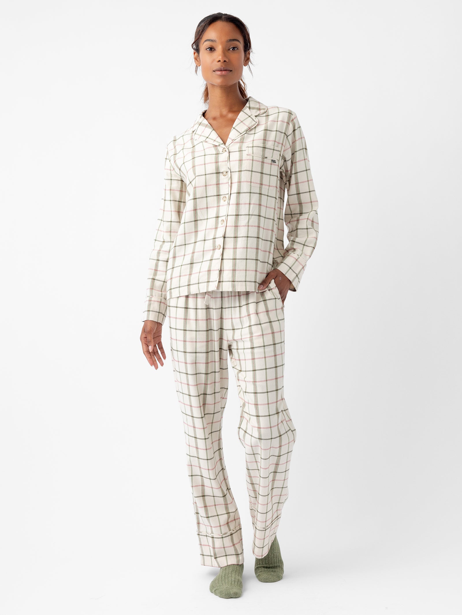 A person wearing Cozy Earth's Women's Farmhouse Long Sleeve Pajama Set, featuring green and pink plaid patterns on a white background, stands against a plain white backdrop. They also have on green socks. The outfit includes a button-up long-sleeved top and matching pants.