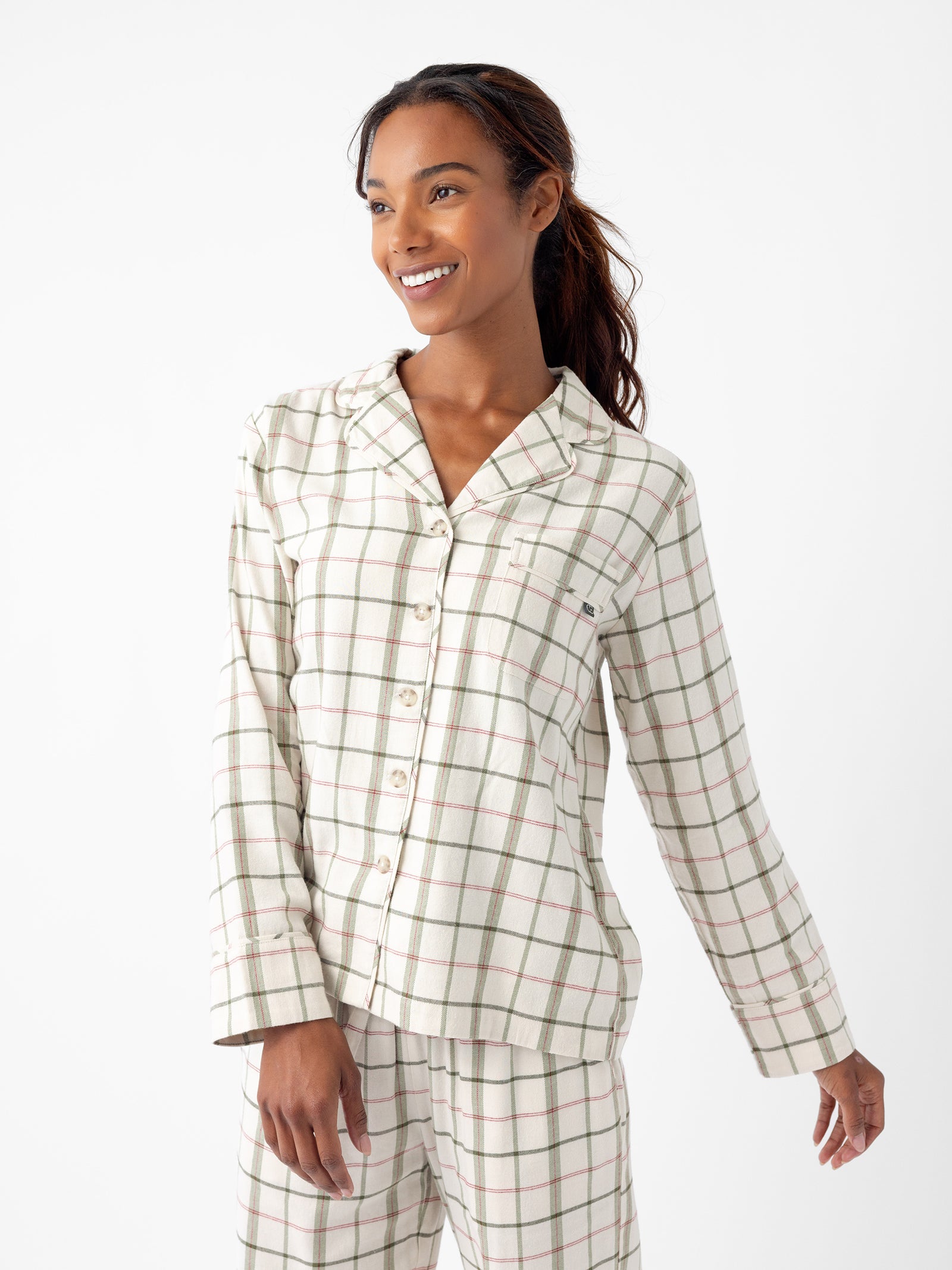 A person with long hair smiles while wearing the Women's Farmhouse Long Sleeve Pajama Set from Cozy Earth. The pajamas feature a green and beige checkered pattern, complete with a button-up shirt that includes a chest pocket and matching pants. The background is white and minimalistic. 