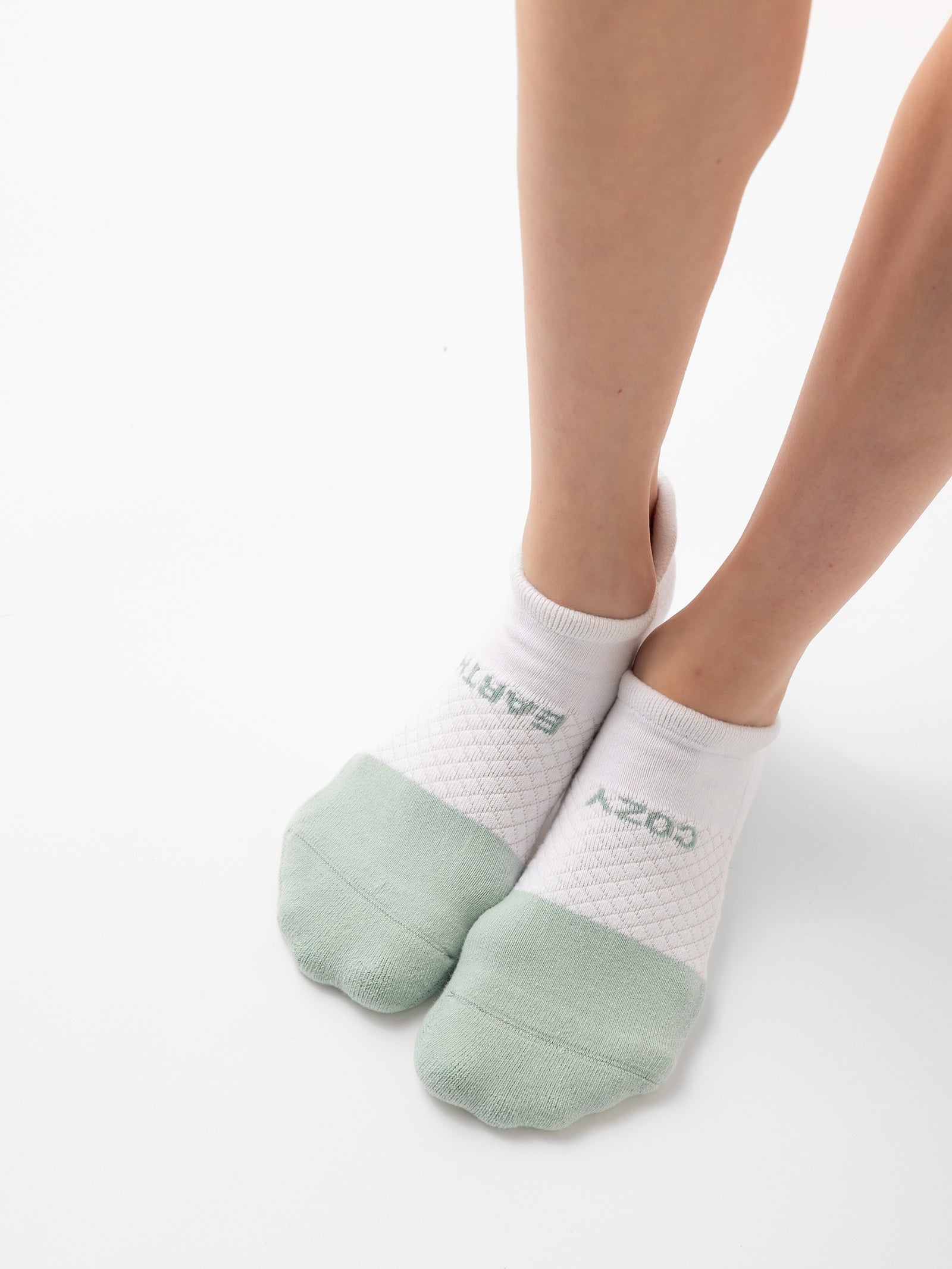 A person is wearing Cozy Earth's Essential Ankle Sock 2-Pack, featuring white ankle socks with a light green toe area and text near the ankles. The person is standing on a white surface with legs slightly bent at the knees, casting a soft shadow below. 