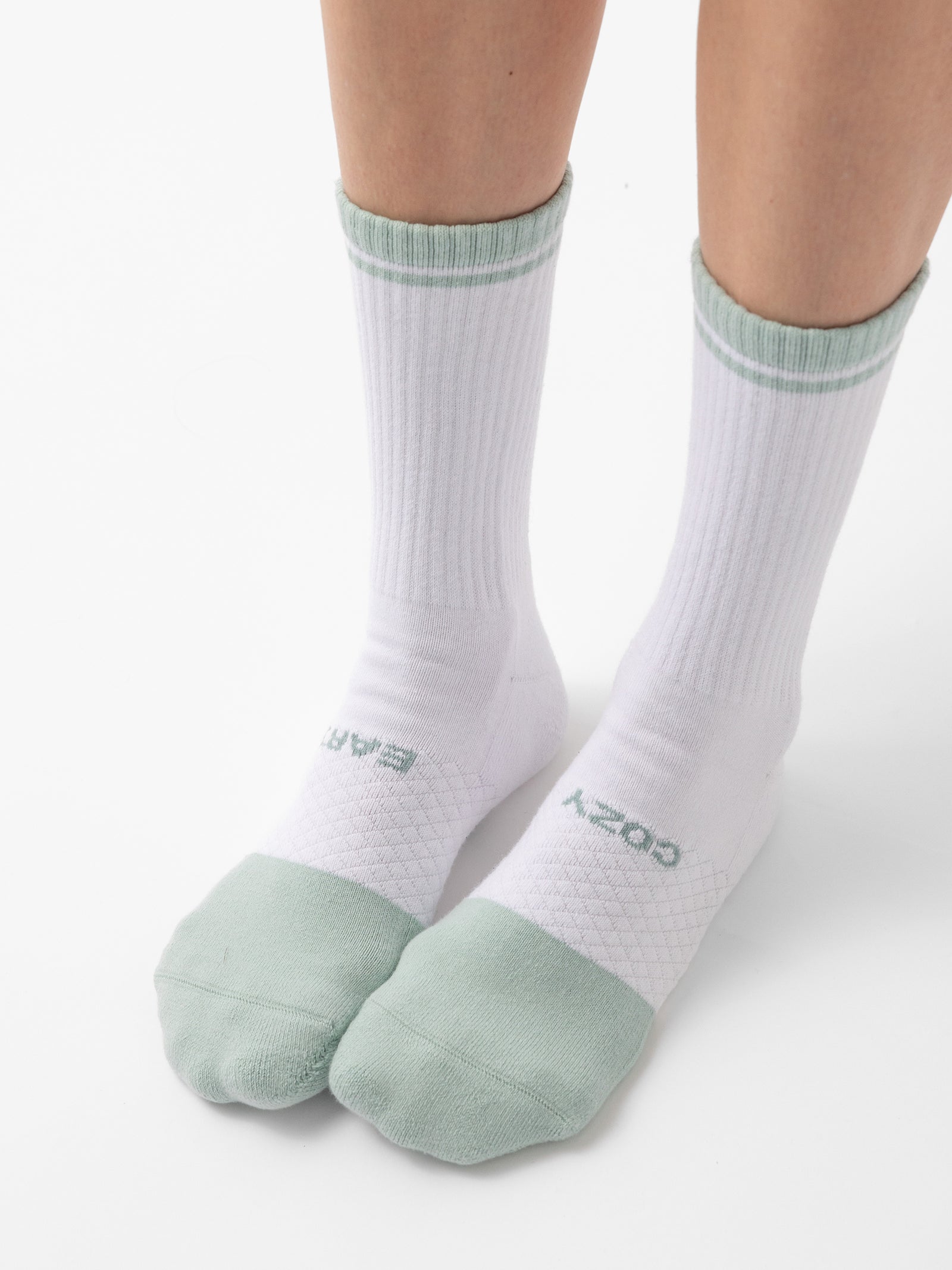 A pair of legs wearing Cozy Earth's Essential Calf Socks in white, featuring light green toes and cuffs. The socks display a ribbed texture extending from the ankles upward and a diamond-knit pattern on the lower foot, with "COZY" and another partially visible word embossed near the toes. 