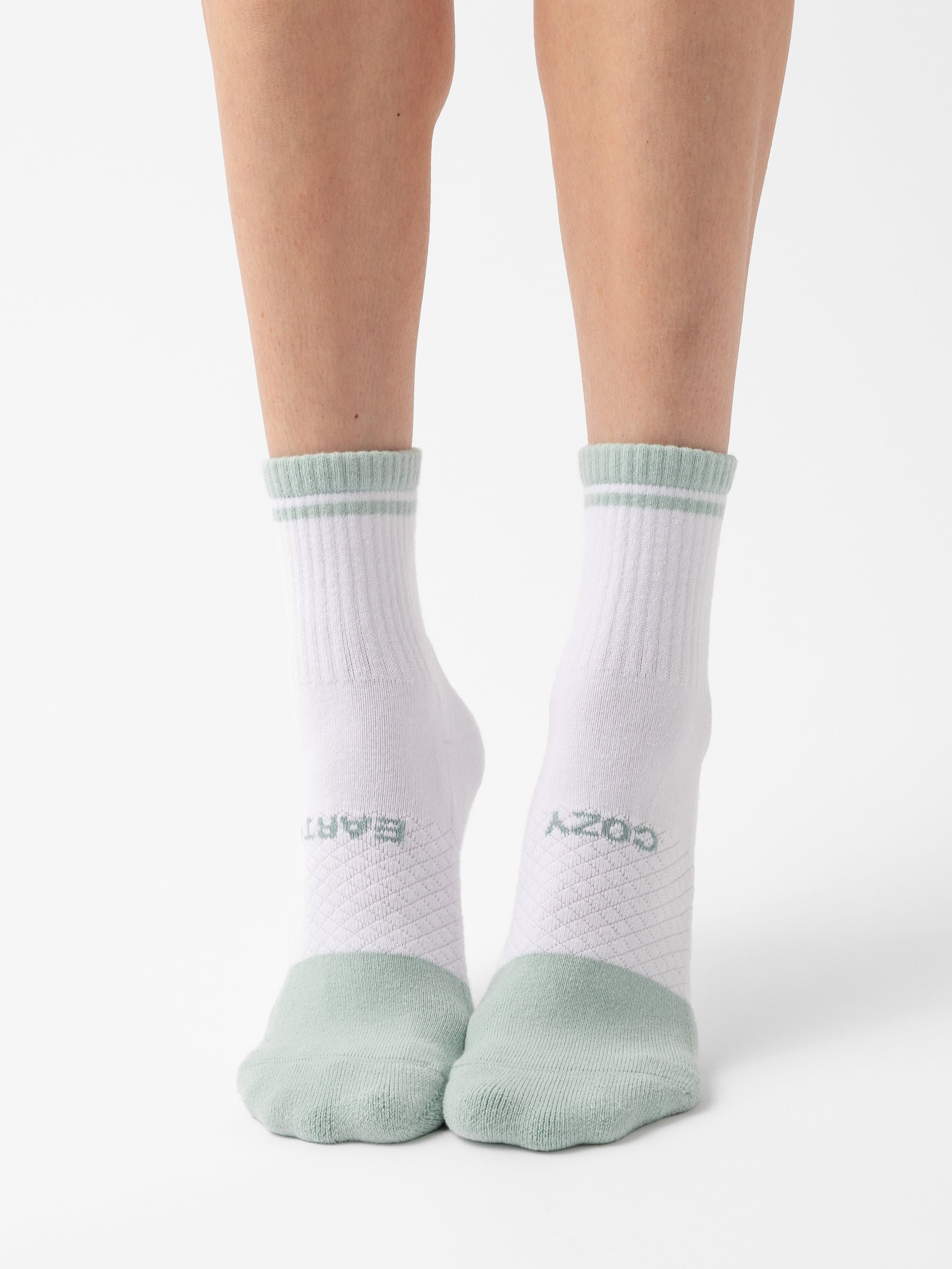 A pair of legs wearing the Essential Quarter Sock 2-Pack by Cozy Earth, featuring white ankle socks with mint green toes, heels, and cuffs. The socks also display text on the top and sole, with "COZY" visible on one sole. The background is plain white. 