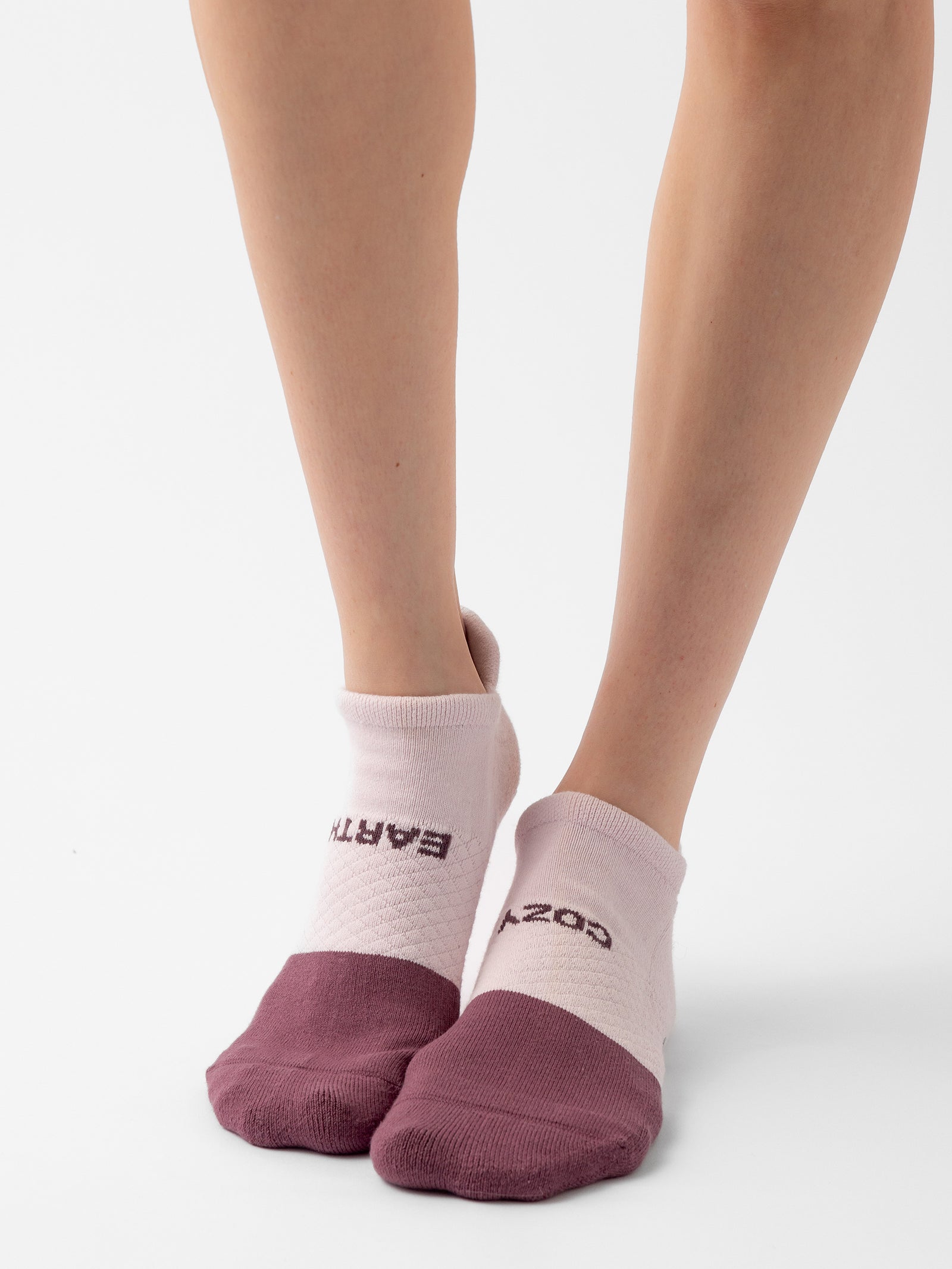 Close-up of a person's lower legs wearing ankle socks from the Essential Ankle Sock 4-Pack by Cozy Earth. The socks feature a two-tone design with a light pink main section and maroon toes, heels, and cuffs. The words "EARTH" and "COZY" are also printed on them. The background is white. 