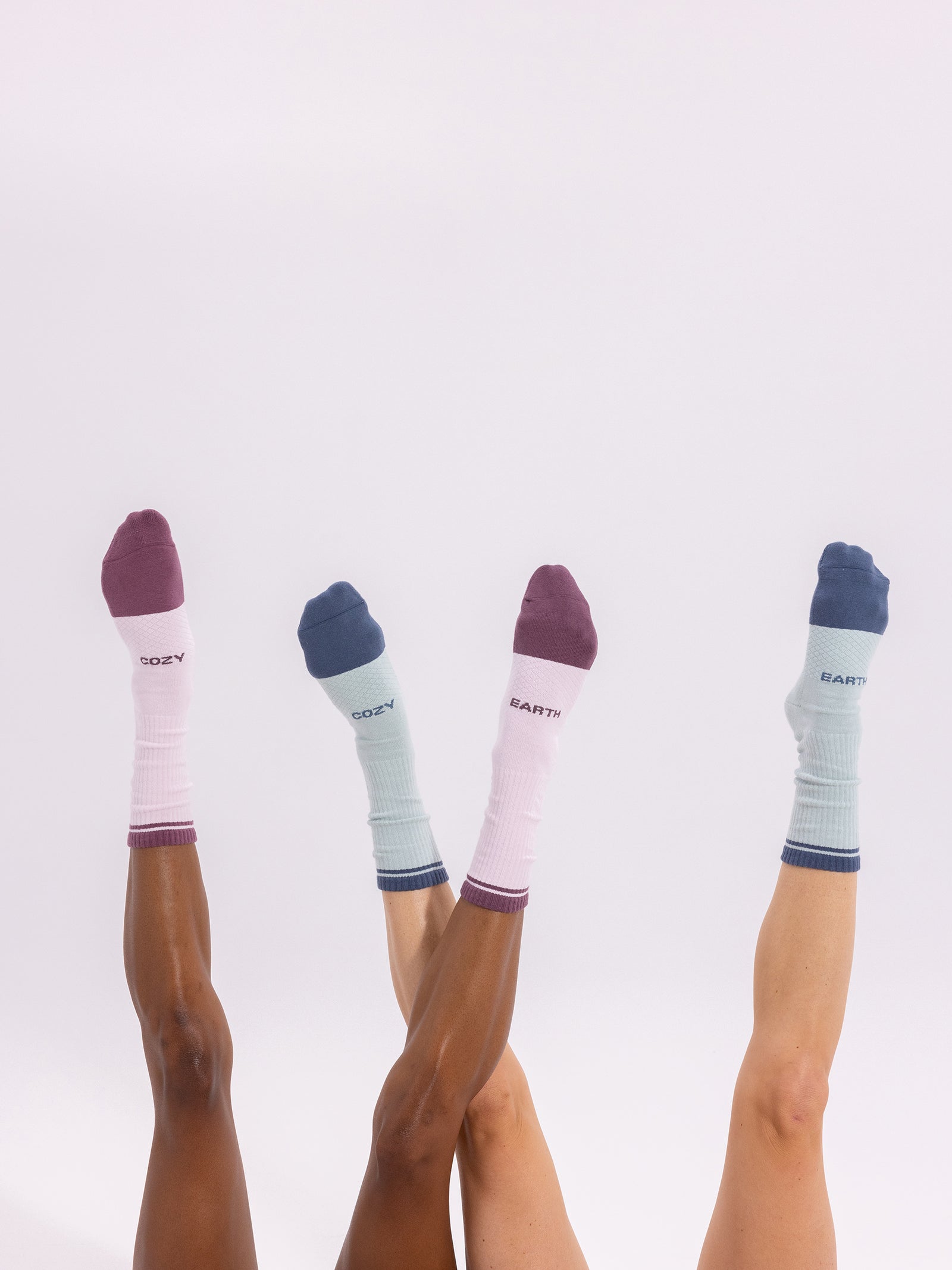 Three pairs of legs in colorful socks from the Cozy Earth Essential Calf Sock 4-Pack are playfully stretched upwards against a white background. The socks come in various pastel shades, with some featuring words like "COZY" and "EARTH" printed near the toes. 