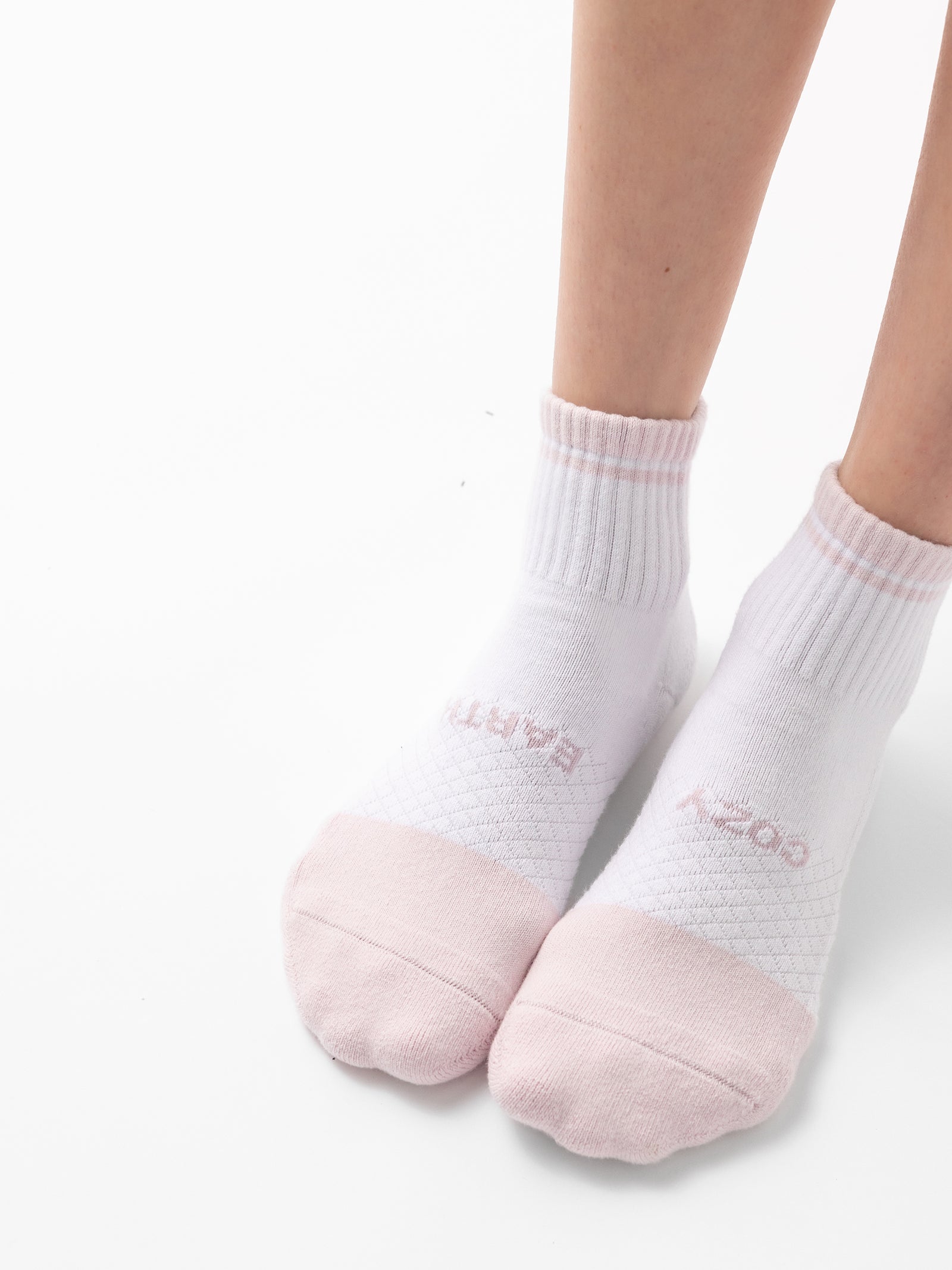 The image showcases feet adorned in Cozy Earth's Essential Quarter Sock from their 4-Pack collection. These white ankle socks are accented with pastel pink toe caps and heel sections. They feature a subtle ribbed texture at the ankle and a delicate, soft pattern around the midsection, all while standing on a white surface. 