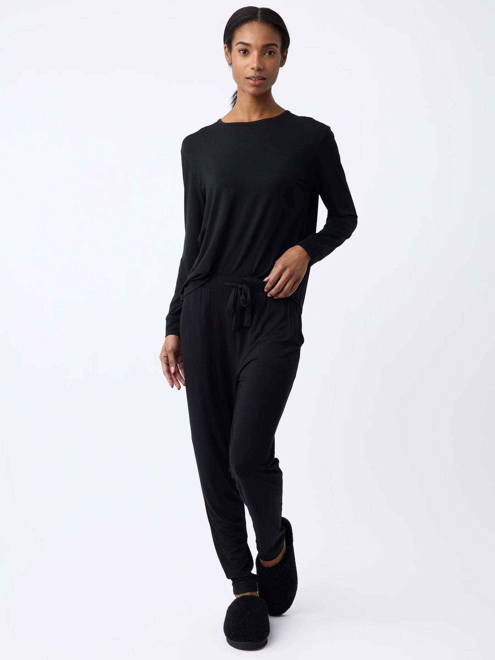 A woman in a Cozy Earth Women's Stretch Knit Bamboo Long Sleeve Lounge Tee and black slipper-like shoes strikes a pose against a white background. Their relaxed-fit jumpsuit with drawstring waist includes one hand resting in their pocket. 