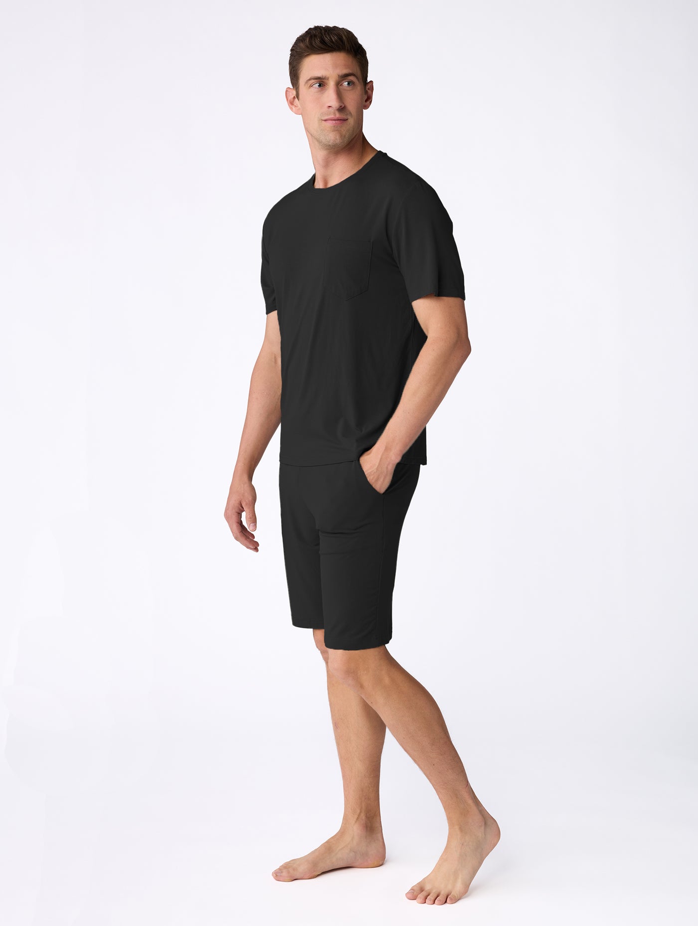 A man stands barefoot on a white background, wearing the Cozy Earth Men's Bamboo Stretch-Knit Short Sleeve Pajama Set in black, featuring a matching short-sleeve T-shirt and pocketed shorts, looking slightly to his left. 