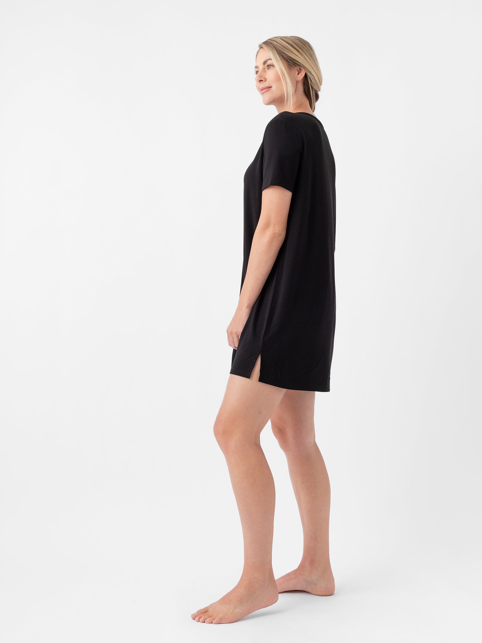 A woman with blonde hair is standing barefoot on a white background. She is wearing Cozy Earth's Women's Bamboo Stretch Knit Sleep Dress, a black, short-sleeved, knee-length dress with a small side slit. She poses with one leg slightly bent and looks off into the distance. 