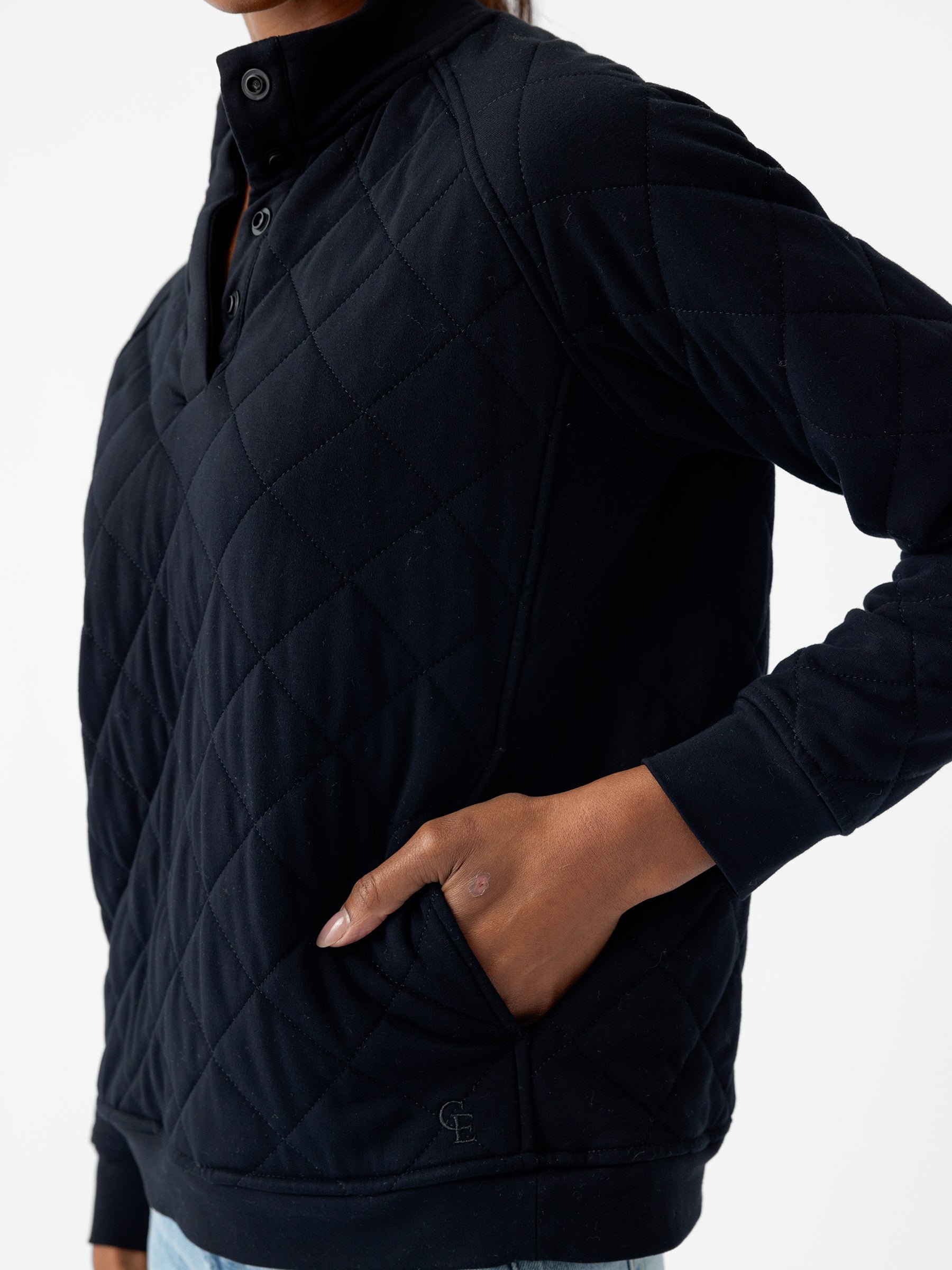 A person is wearing a Women's Ultra-Soft Bamboo Quilted Snap Pullover from Cozy Earth, featuring a high collar and buttons. They have one hand in their front pocket and are standing against a plain white background, with the initials "GE" embroidered near the pocket. 