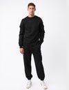 A man stands against a white background wearing the Men's CityScape Crewneck & Sweatpant Set by Cozy Earth in black, paired with white sneakers. He faces forward with one hand in his pocket and a neutral expression.|Color:Black
