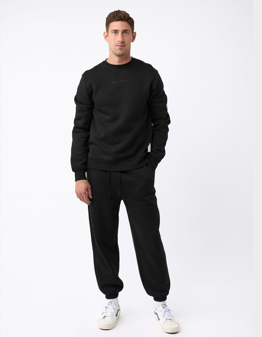 A man stands against a white background wearing the Men's CityScape Crewneck & Sweatpant Set by Cozy Earth in black, paired with white sneakers. He faces forward with one hand in his pocket and a neutral expression.|Color:Black
