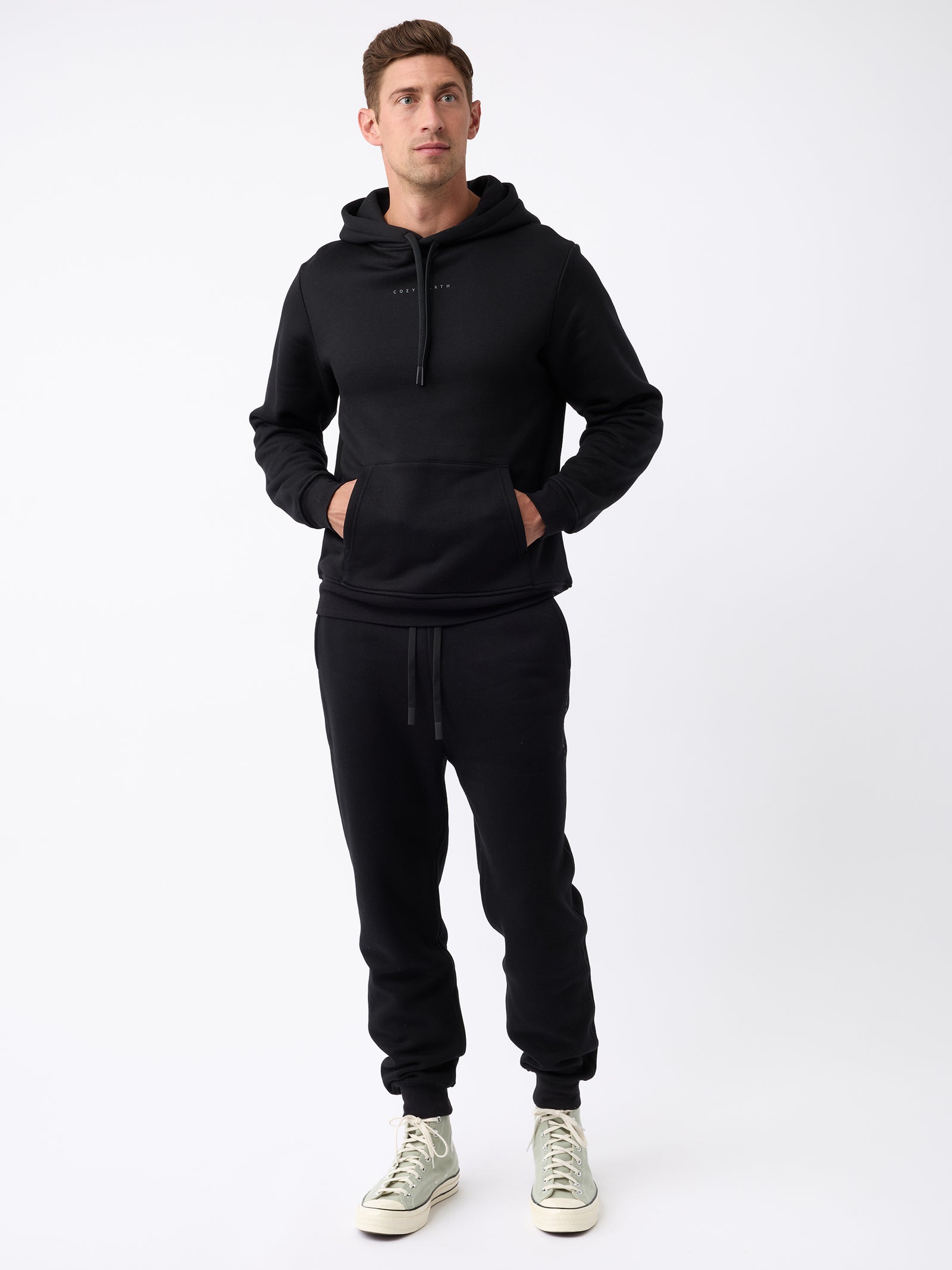 An individual is wearing the Cozy Earth Men's CityScape Jogger in black, along with a black hoodie. Their hands are tucked into the hoodie pocket, and they stand against a white background. They are sporting light-colored high-top sneakers. 