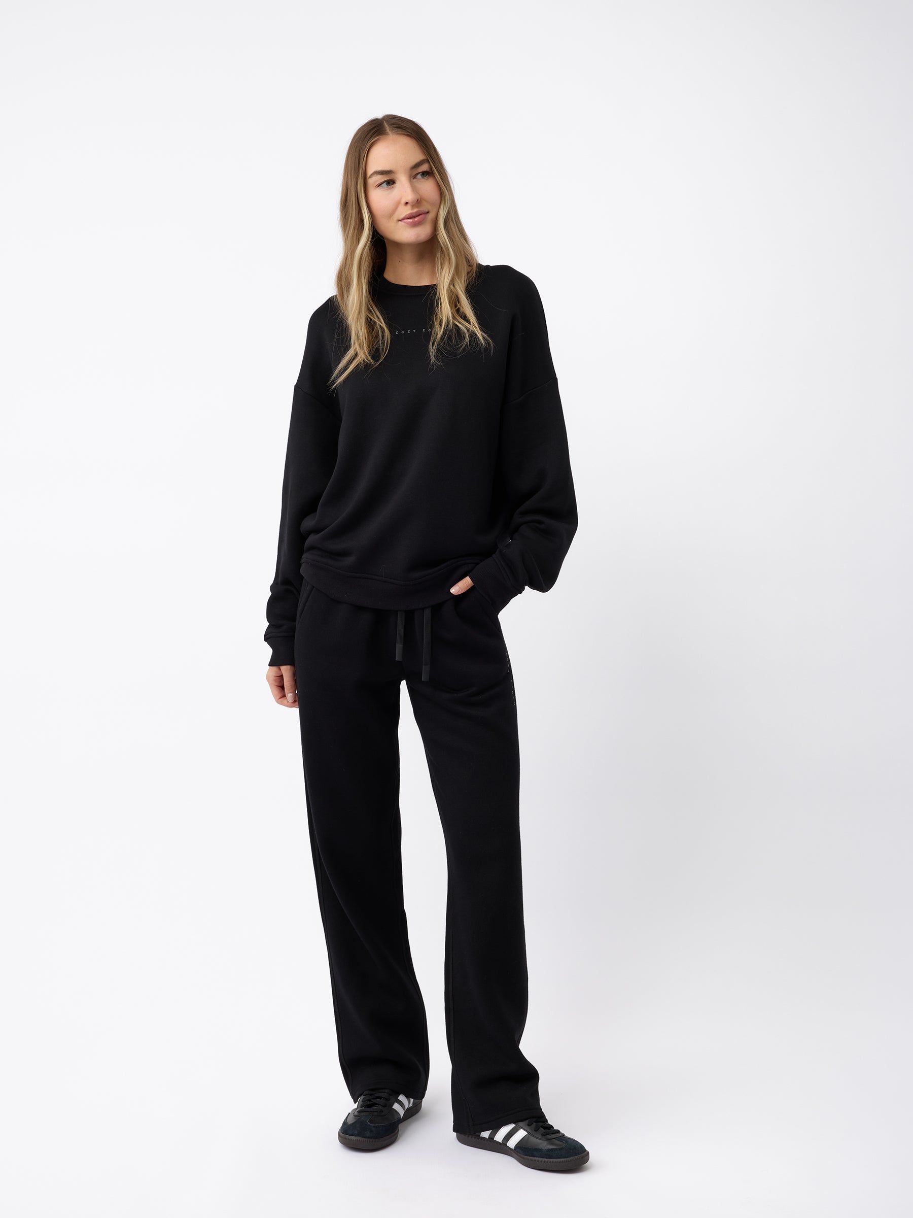 A person with long hair stands against a plain background, dressed in a black sweater and Cozy Earth's Women's CityScape Wide Leg Pant, with their left hand in a pocket. They are also wearing black and white sneakers. |Color:Black