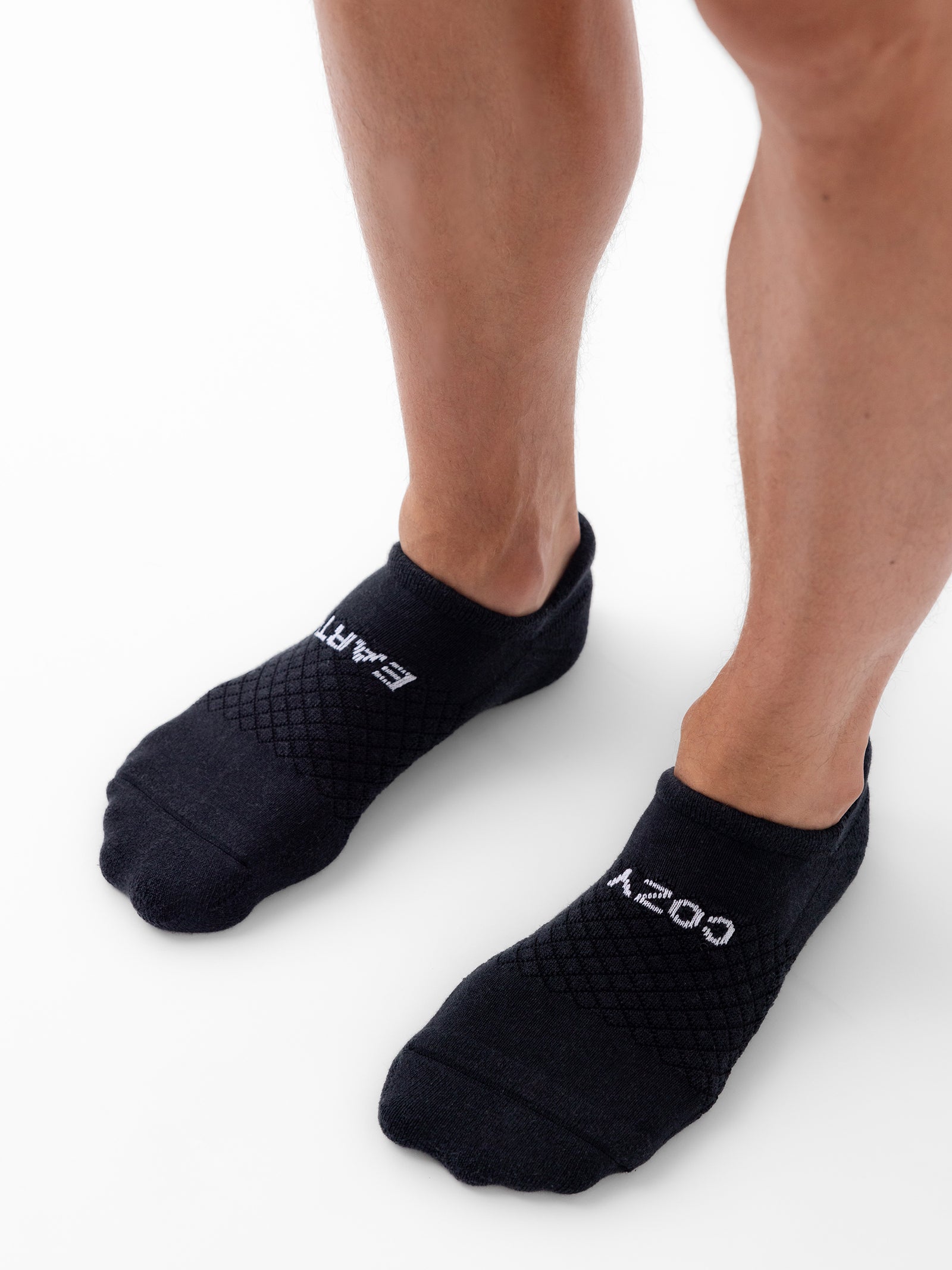 A close-up of a person's legs and feet wearing black socks from the Essential Ankle Sock 2-Pack by Cozy Earth. The socks showcase the text "COZY" on the left sock and "EARTH" on the right sock. The shot features the person's legs from the knees down, standing on a plain white background. 