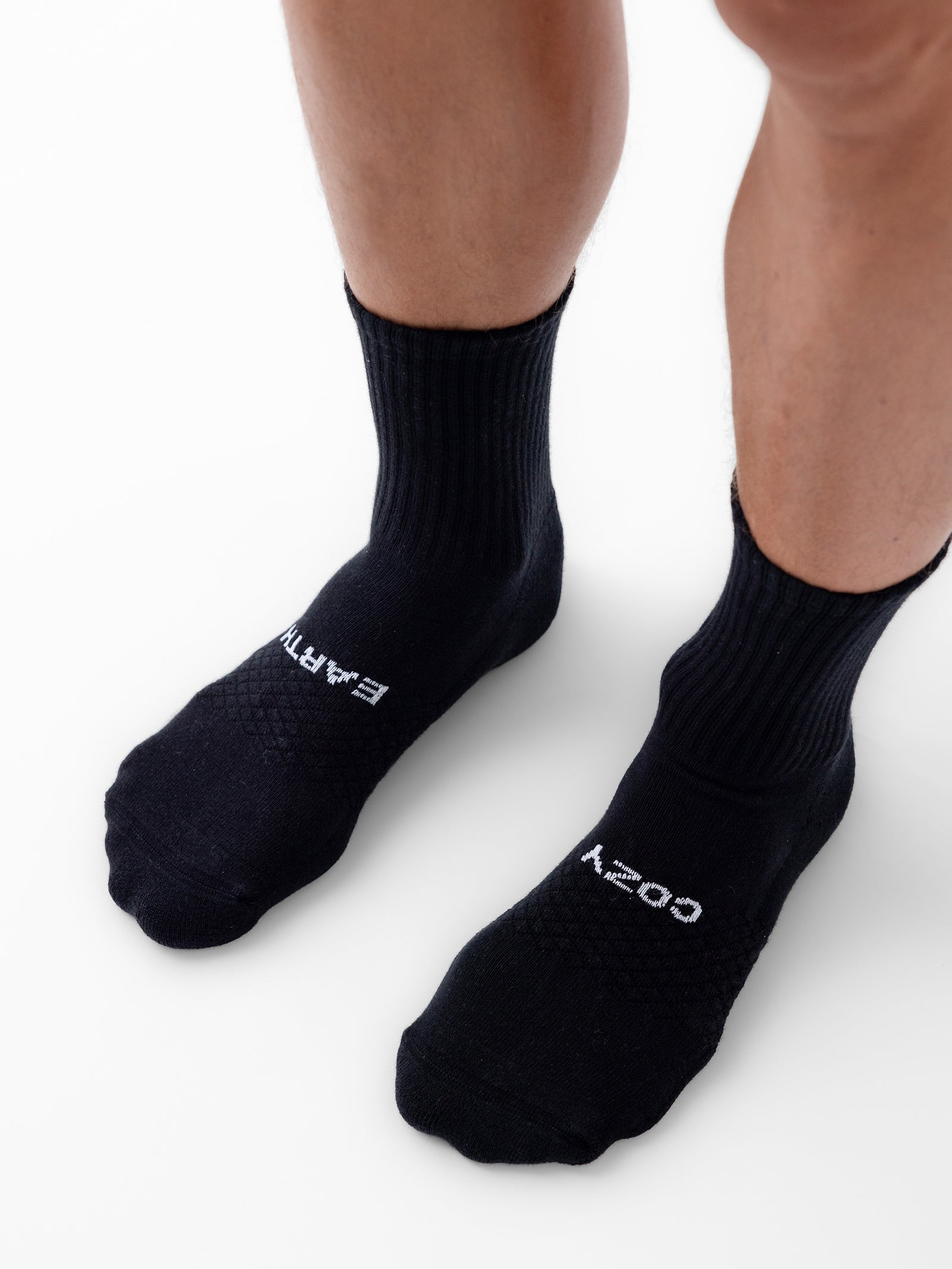 A pair of legs wearing black socks from the Cozy Earth Essential Calf Sock 4-Pack, with "EARTH" written on the left sock and "COZY" written on the right sock in white text. The person is standing on a clean, white surface. 