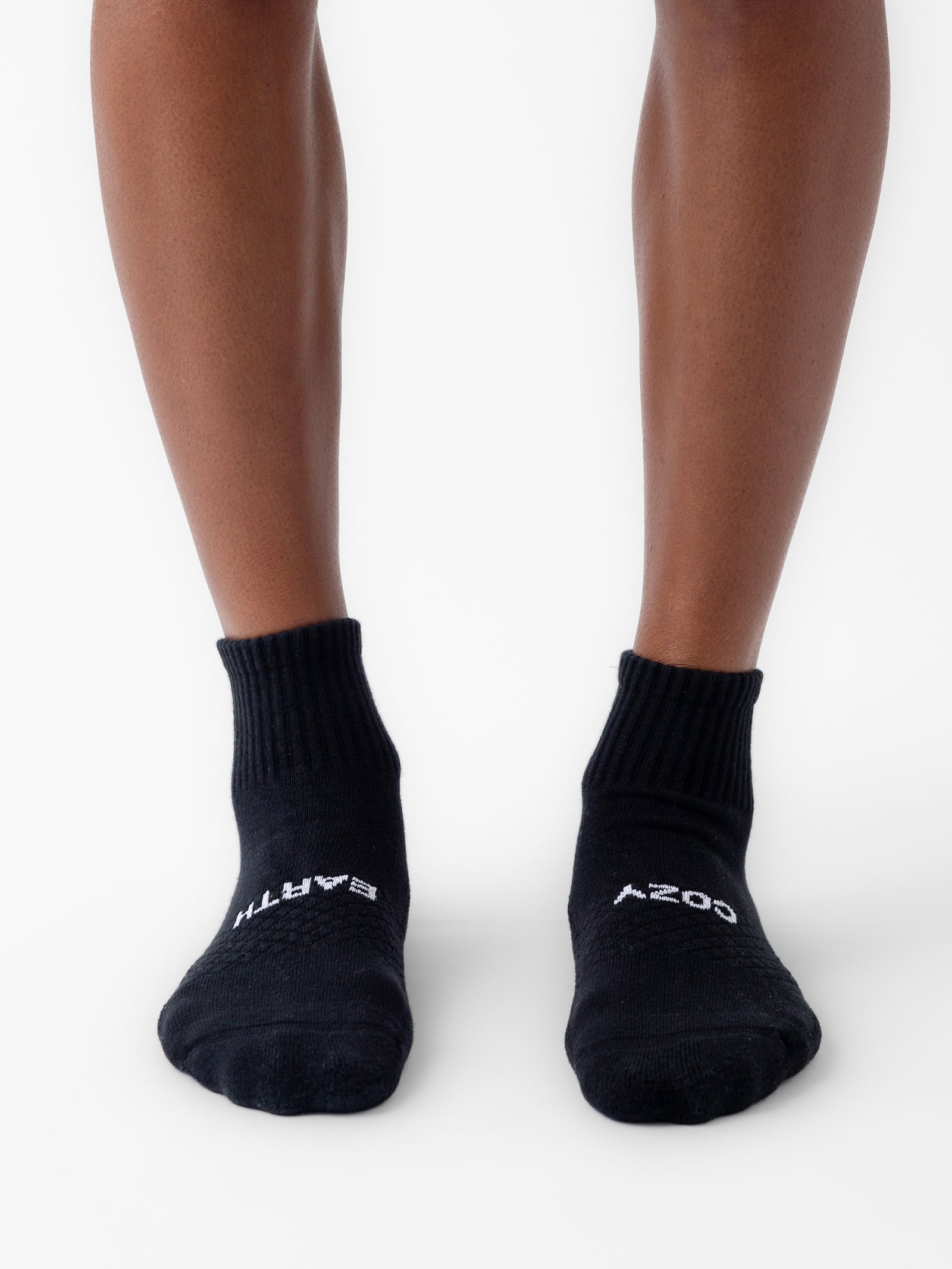 A person stands with feet slightly apart, wearing black ankle socks from the Essential Quarter Sock 2-Pack by Cozy Earth. The socks feature white text—"EARTH" on the left sock and "COSY" on the right sock. The background is plain white. 