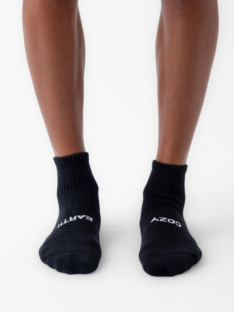 A person wears black Cozy Earth Essential Quarter Socks on a white surface. The socks have white text: "EARTH" on the left toe and "COZY" on the right.