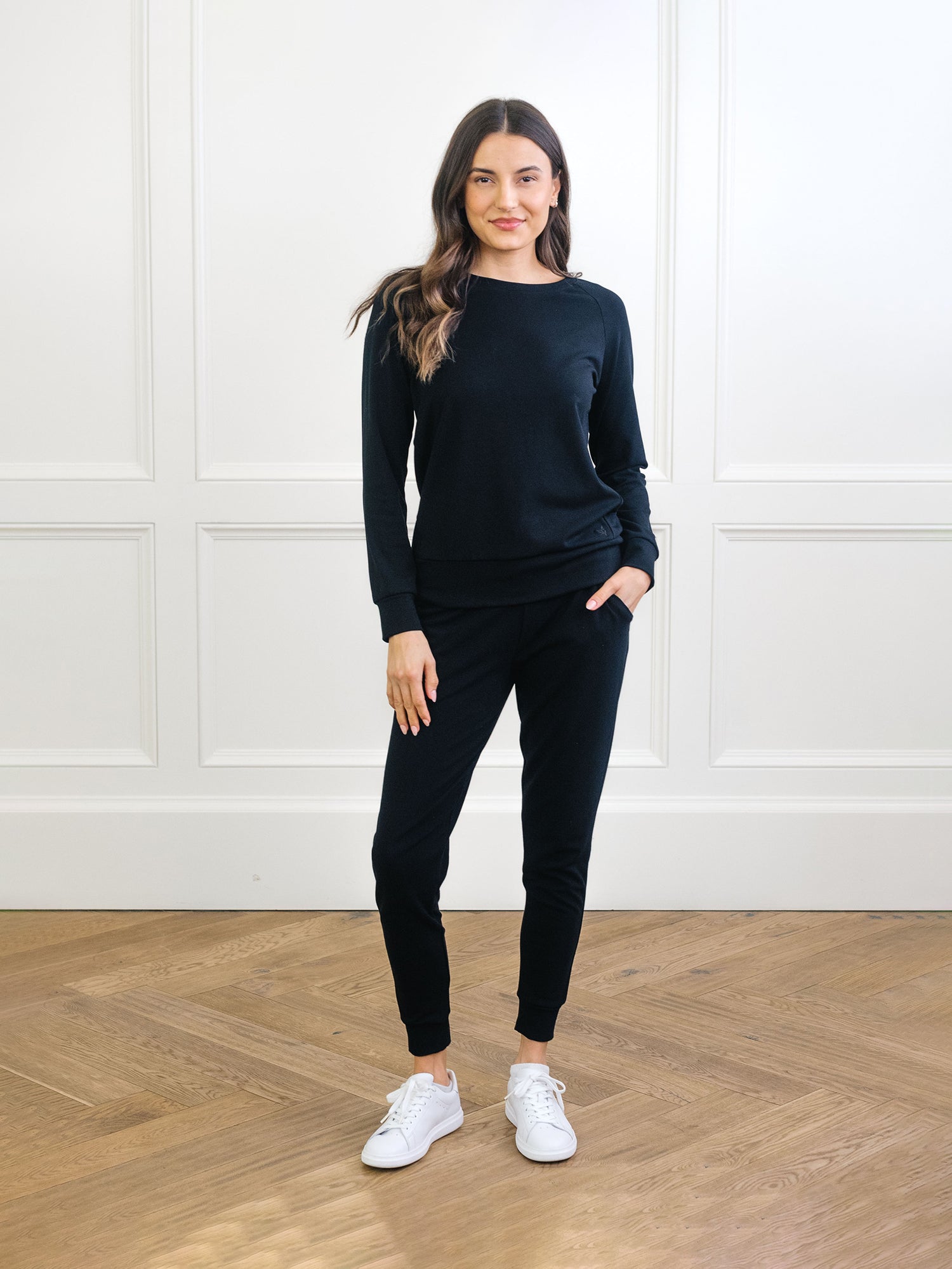 A person with long hair stands against a white-paneled wall, wearing Cozy Earth's Women's Bamboo Jogger Set in black. They pair it with white sneakers and pose confidently on a wooden floor, one hand in their pocket. |Color:Black