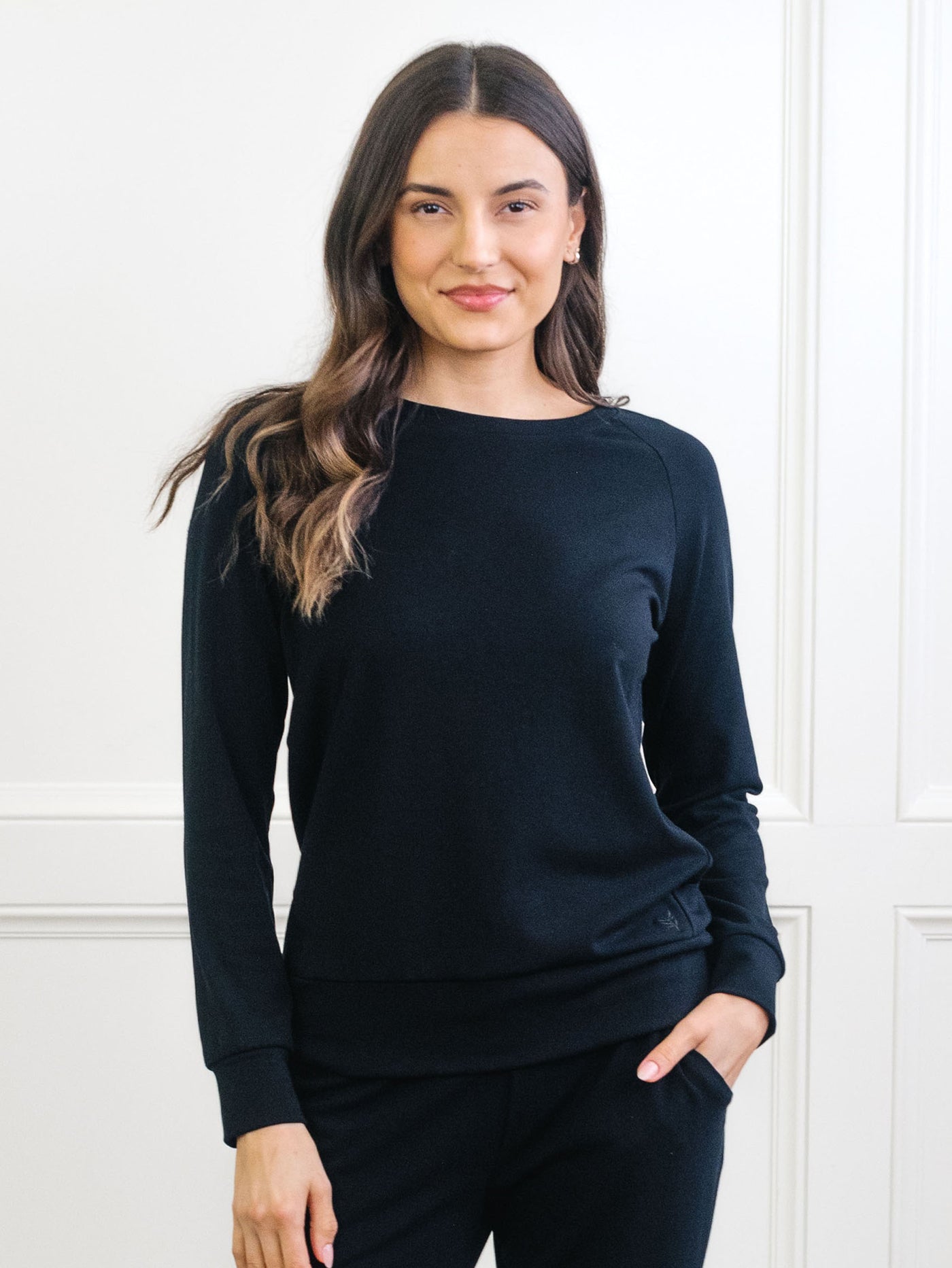 A woman with long brown hair smiles with one hand in her pocket, wearing a Cozy Earth Women's Ultra-Soft Bamboo Wide Leg Pull On Pant & Pullover Crew Set against a white paneled wall. 