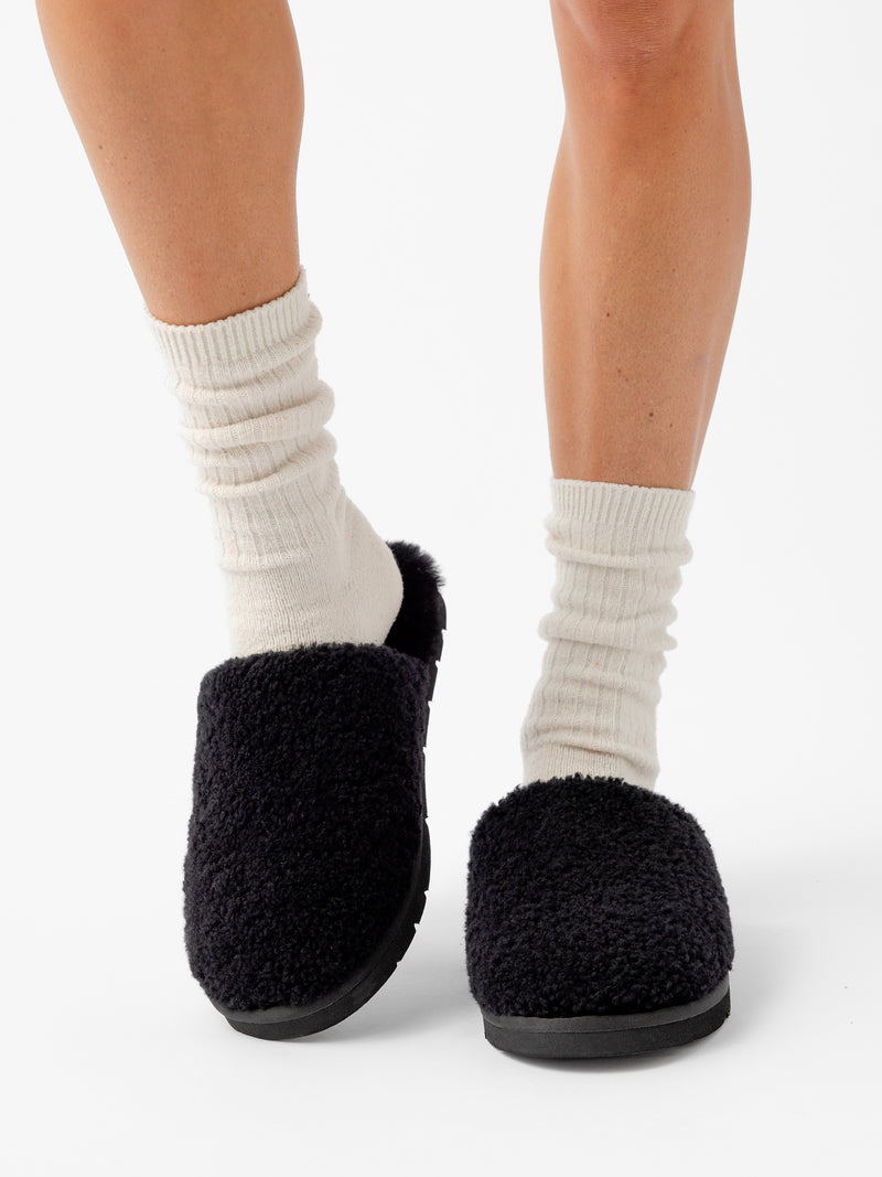 A person wearing white, ribbed socks and Cozy Earth's Puffy Sheep Slippers is standing on a white background. Only their lower legs and feet are visible. The slippers have a cozy, textured appearance that contrasts with the light-colored socks.