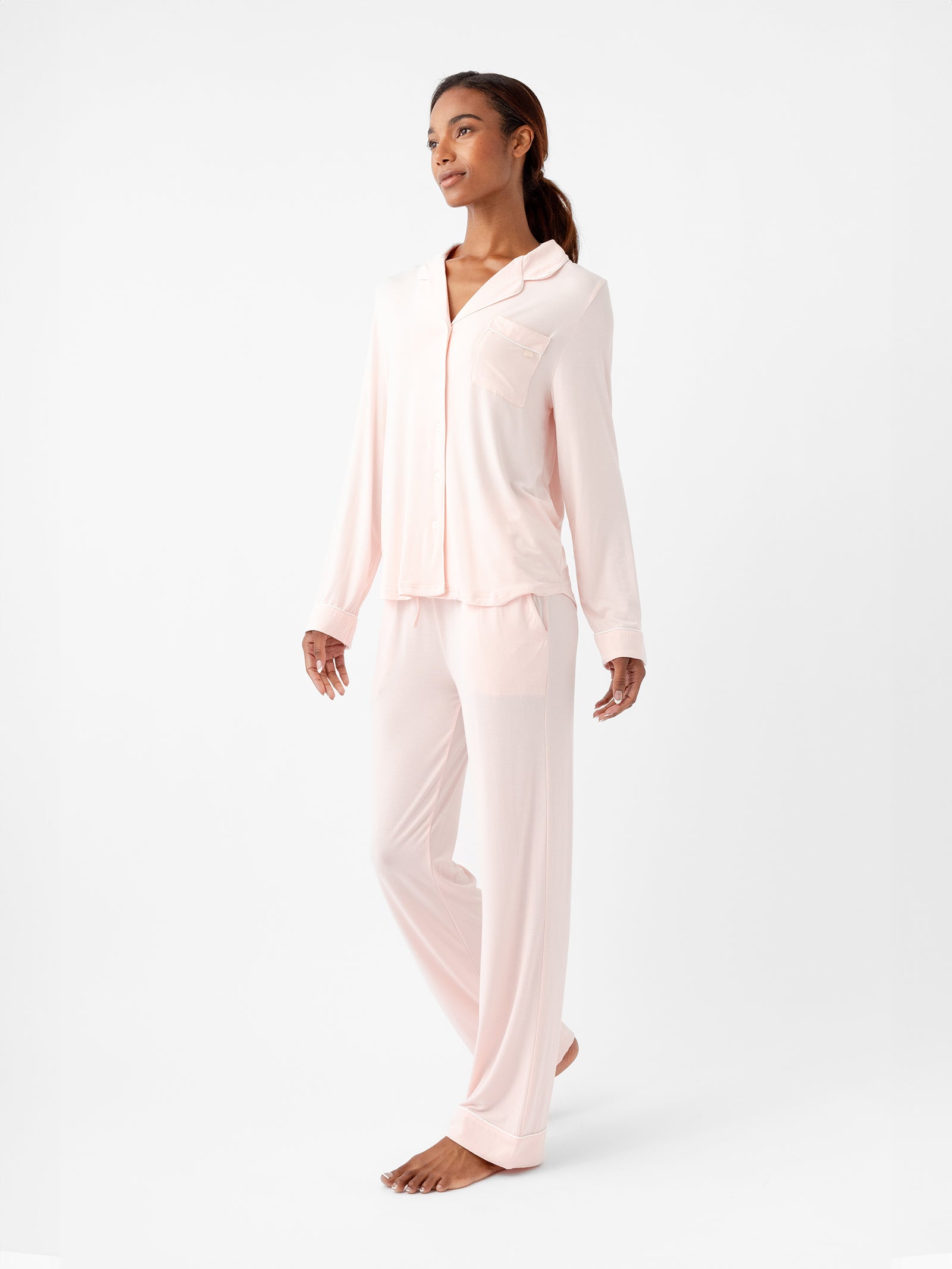 A person is wearing the Women's Long Sleeve Bamboo Pajama Top in Stretch-Knit TALL by HIDE in light pink and loose-fitting matching pants. They are standing barefoot on a white background, looking off to the side. 