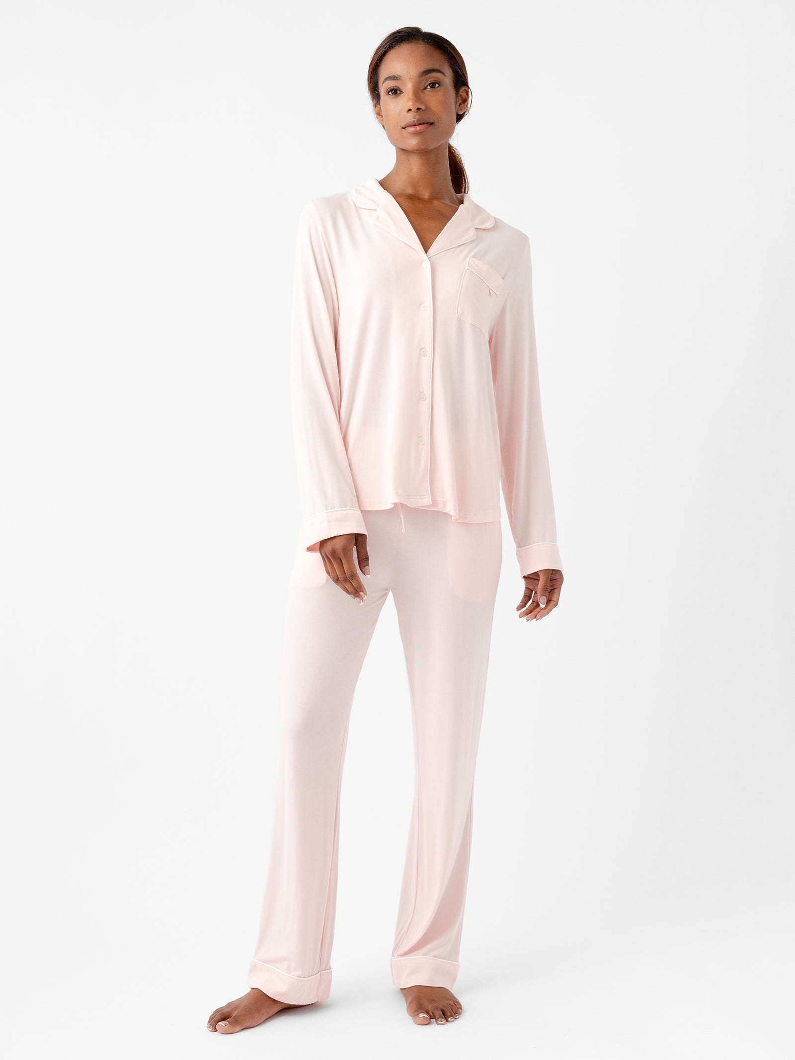 A woman is standing against a plain white background, wearing HIDE's light pink Women's Stretch-Knit Bamboo Pajama Pant set, which consists of a button-up long-sleeve top with a collar and matching pants. She is barefoot and looking slightly off to the side with a relaxed expression. 