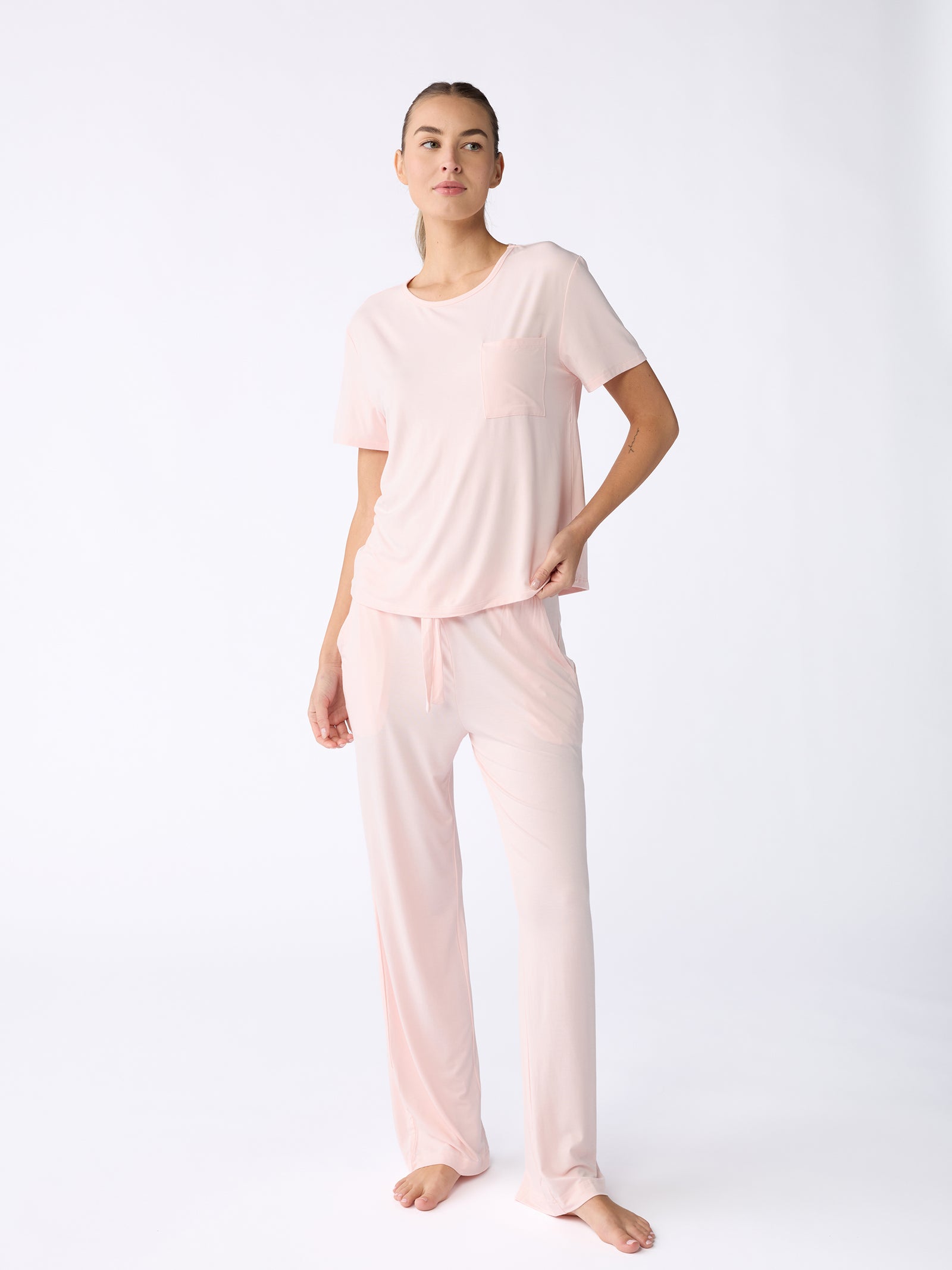 A person is standing barefoot against a plain white background, wearing the Stretch Knit Bamboo Pant from Cozy Earth in light pink. The pajama set includes a short-sleeved top with a chest pocket and pants that feature a relaxed fit. 