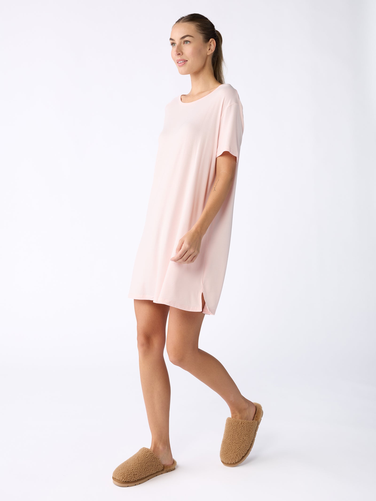 A woman in a pale pink Women's Bamboo Stretch Knit Sleep Dress and brown slippers by Cozy Earth stands against a plain white background. She is smiling, her hair tied back, and the dress is loose-fitting with a casual style. 