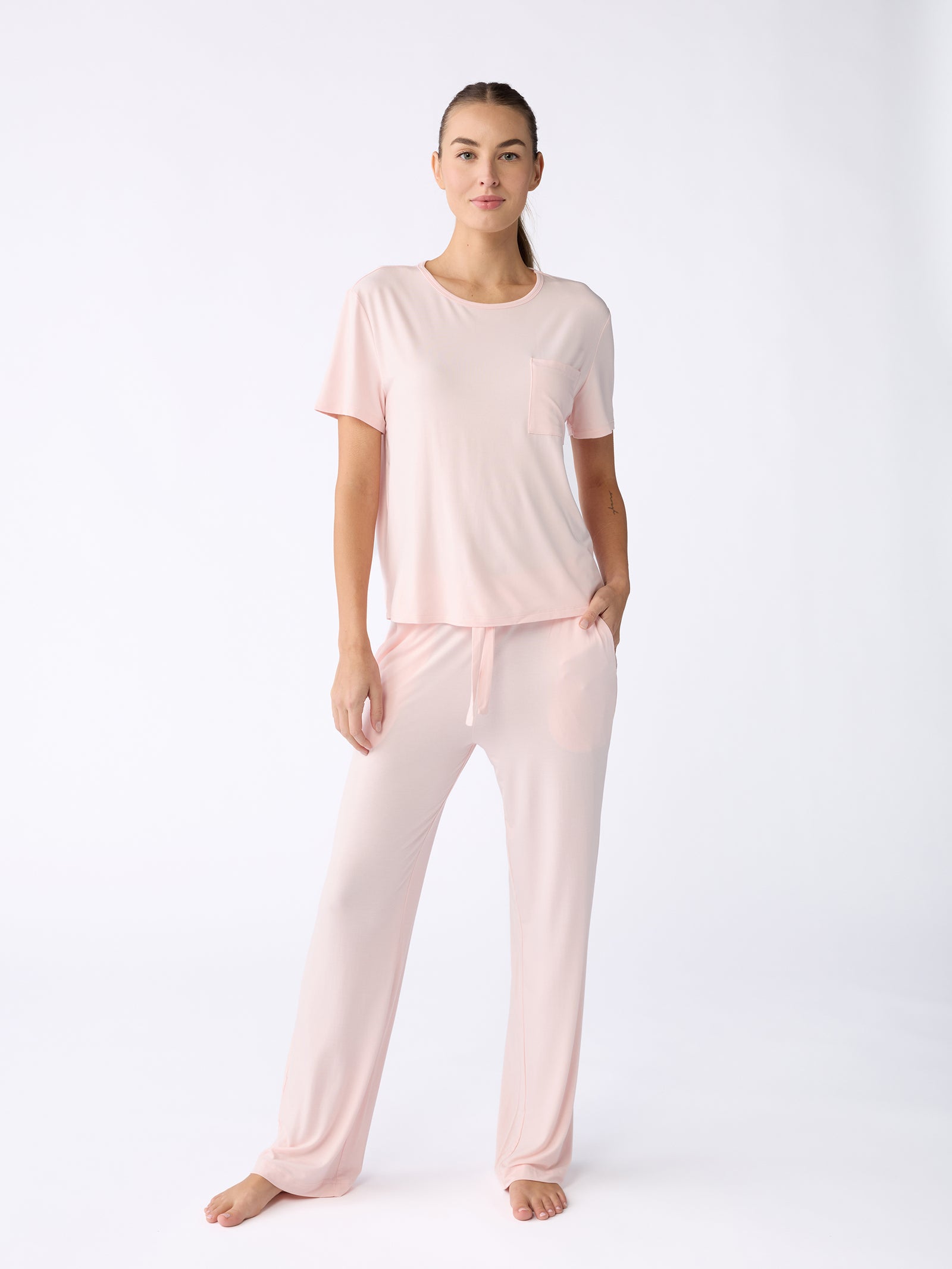 A person stands barefoot against a plain white background, wearing the Women's Stretch-Knit Bamboo Lounge Tee by Cozy Earth—a light pink pajama set featuring a short-sleeve top and long pants. The individual has one hand in a pocket and gazes directly at the camera. 