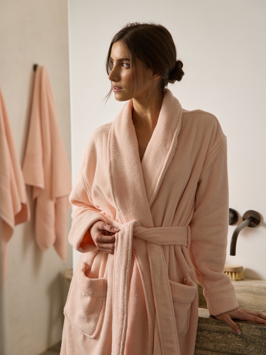 A person stands in a bathroom wearing a Luxe Bath Robe by Cozy Earth, looking to the side. Matching pink towels hang on the wall, and a sink with a faucet is visible beside them, creating an ambiance of relaxation and comfort. 