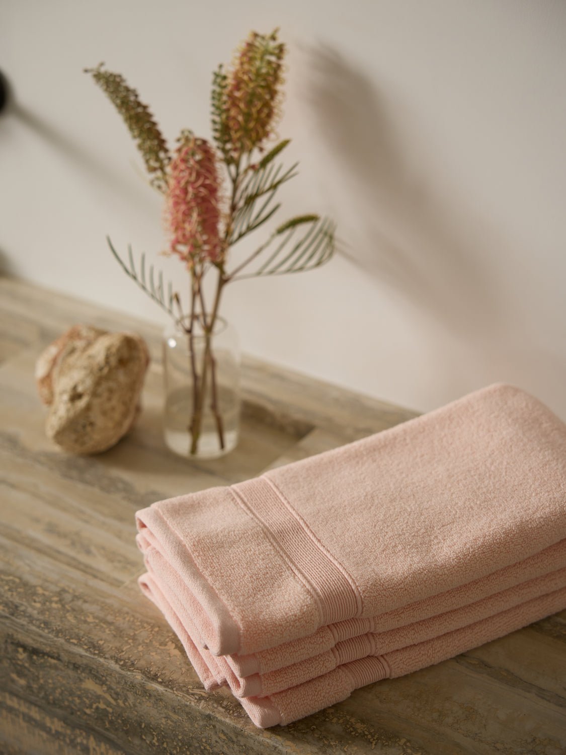 Luxe Hand Towels from Cozy Earth are neatly folded on a wooden surface beside a glass vase with pink flowers and coral. Soft lighting creates a warm, inviting atmosphere. 