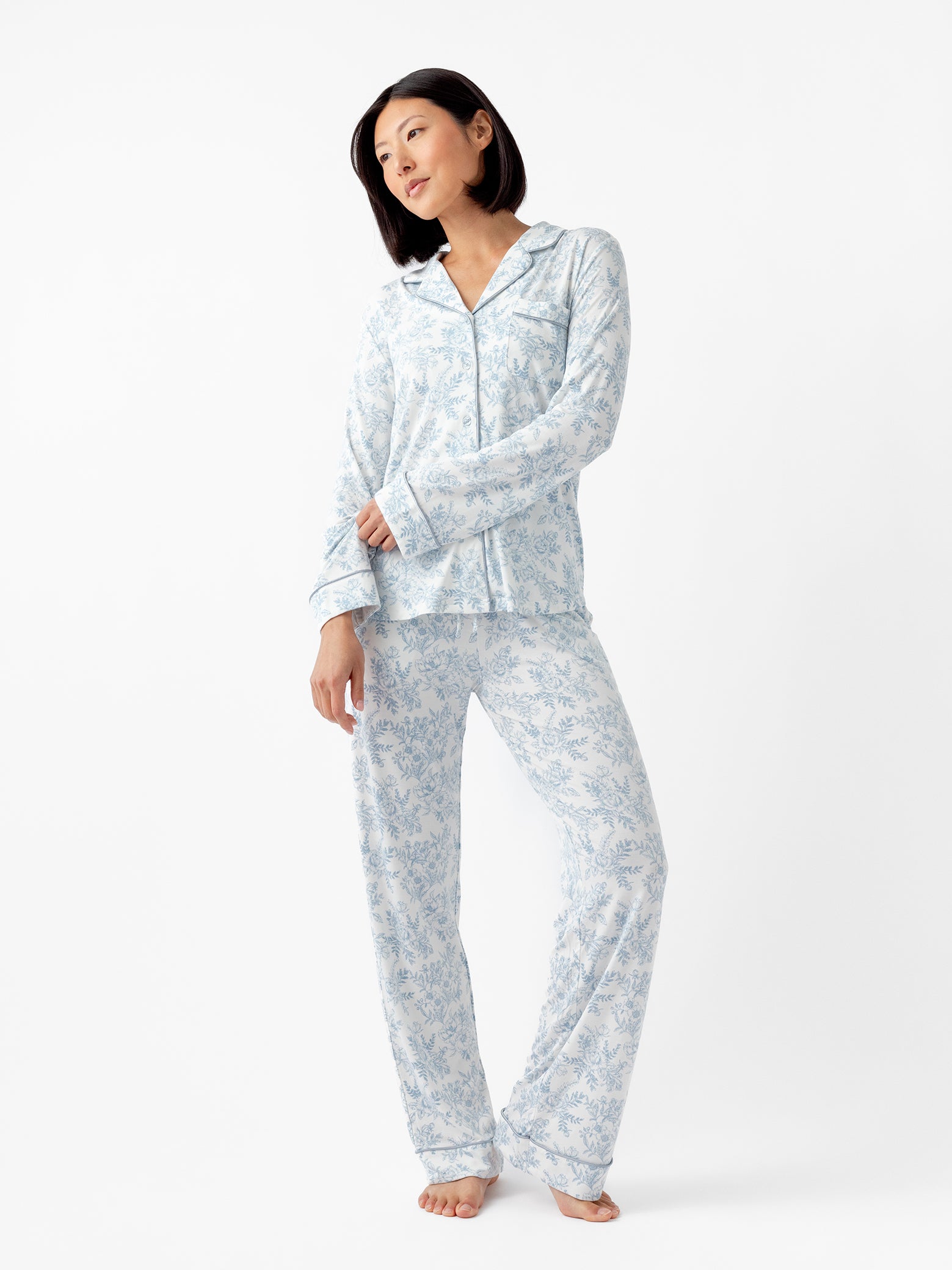 A person models an Cozy Earth Women's Bamboo Stretch-Knit Long Sleeve Pajama Top with light blue floral patterns on a white background, paired with matching pants. They are barefoot and look slightly to the side. 