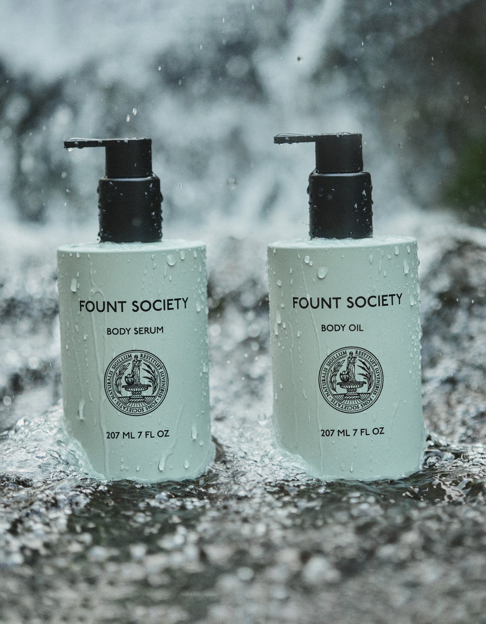 Two pump bottles from the Body Glow Duo by Cozy Earth stand side by side on wet rocks, with water splashing behind them. The left bottle is labeled "Body Serum" and the right bottle is labeled "Body Oil." Both containers have a capacity of 207 ml (7 fl oz) and are adorned with a circular emblem.