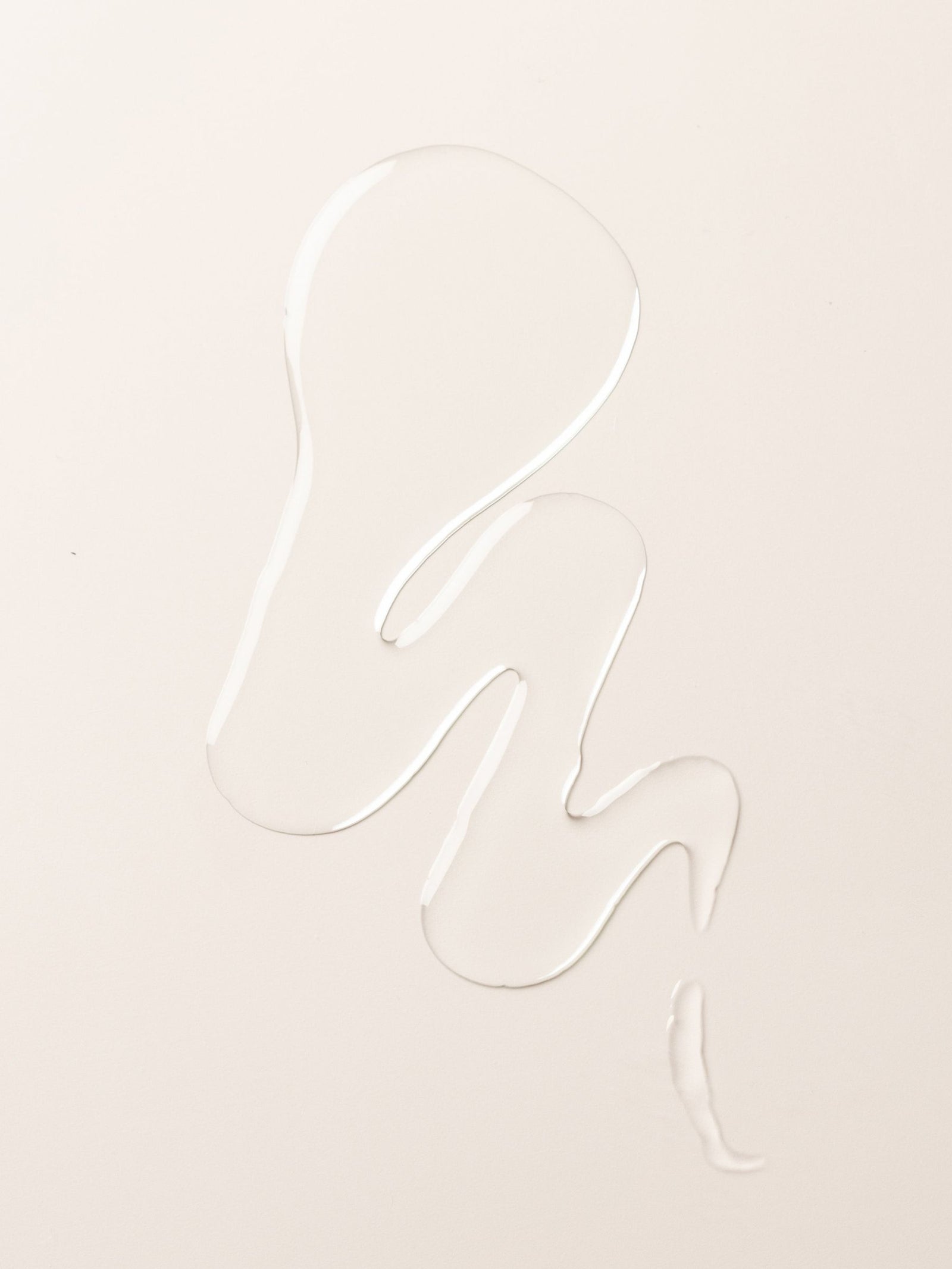 A clear liquid body oil from Cozy Earth forms an abstract, wavy shape on a smooth, off-white surface.