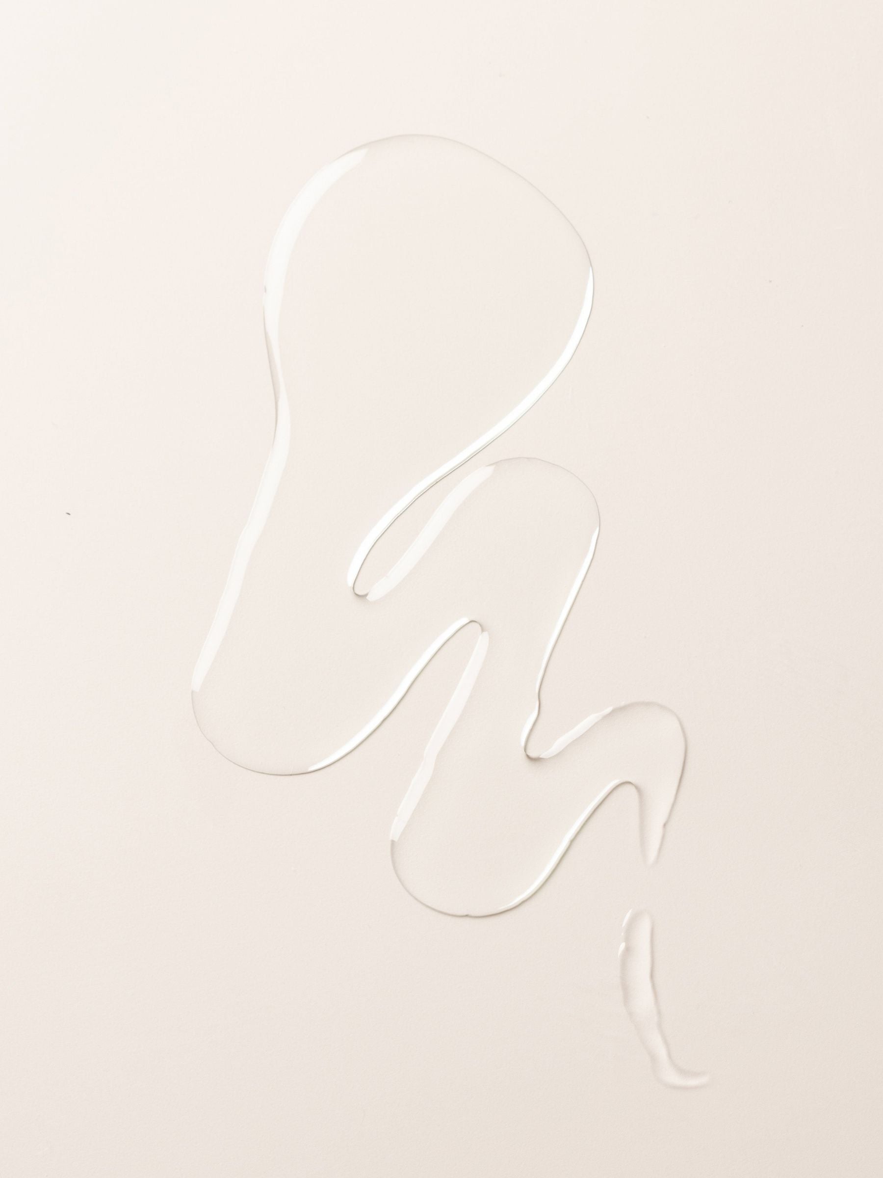 A clear liquid body oil from Cozy Earth forms an abstract, wavy shape on a smooth, off-white surface.