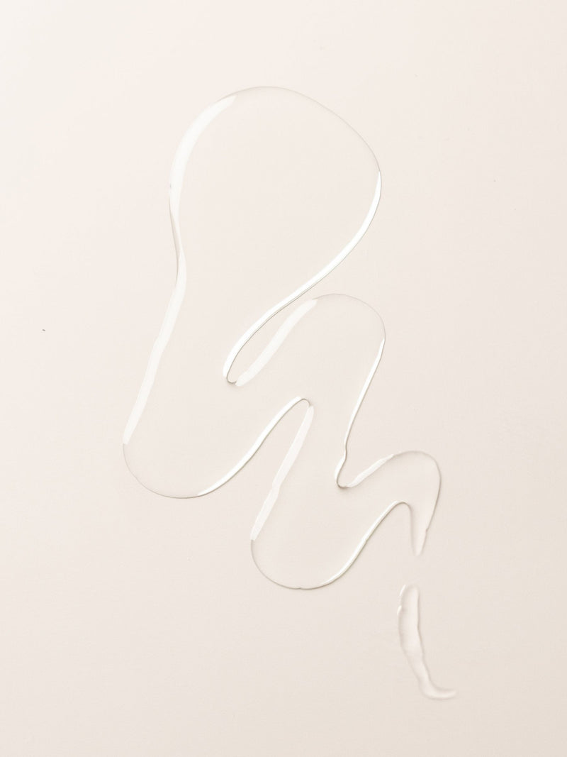 A clear liquid body oil from Cozy Earth forms an abstract, wavy shape on a smooth, off-white surface.