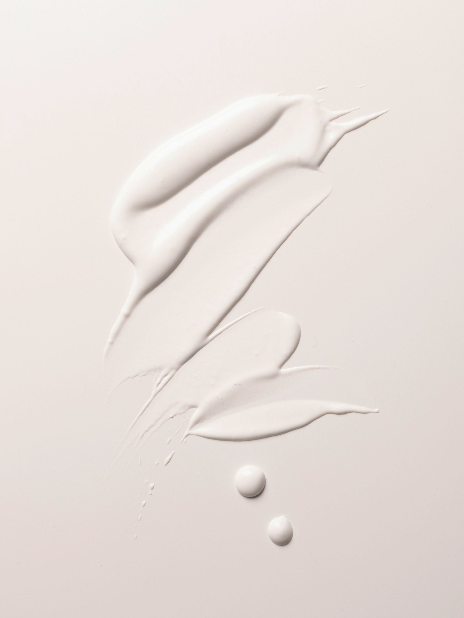 A close-up image featuring Cozy Earth's Body Serum spread on a smooth, light beige surface. The creamy white serum is smeared in an abstract pattern with two small droplets below the main smears.