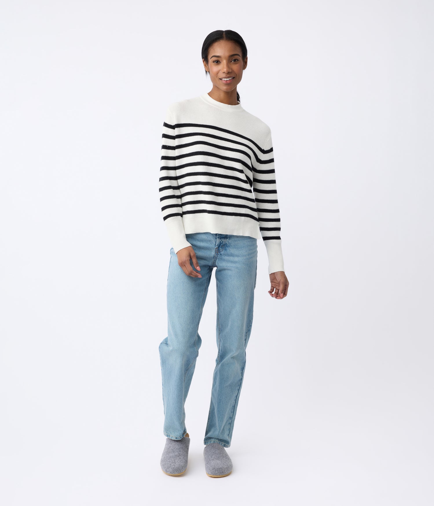 A person stands smiling in a Cozy Earth Women's Rowan Sweater, light blue jeans, and gray slippers against a plain white background. |Color:Bone/Jet Black Stripe