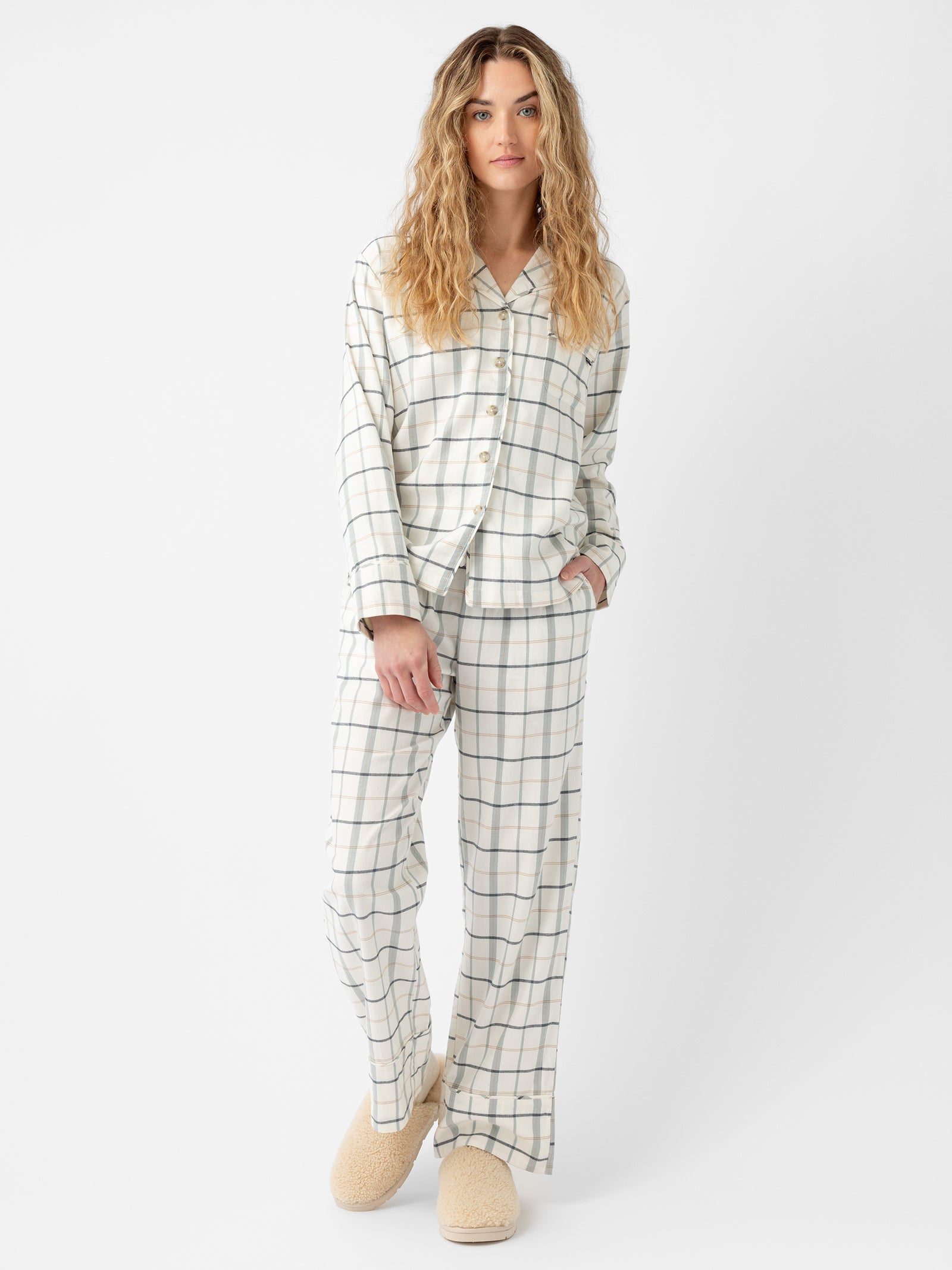 A person with long wavy blonde hair stands against a plain white background, wearing the Cozy Earth Women's Farmhouse Long Sleeve Pajama Set, featuring a light base color with thin plaid lines. The pajama top has buttons, and the person is also wearing cozy beige slippers. 