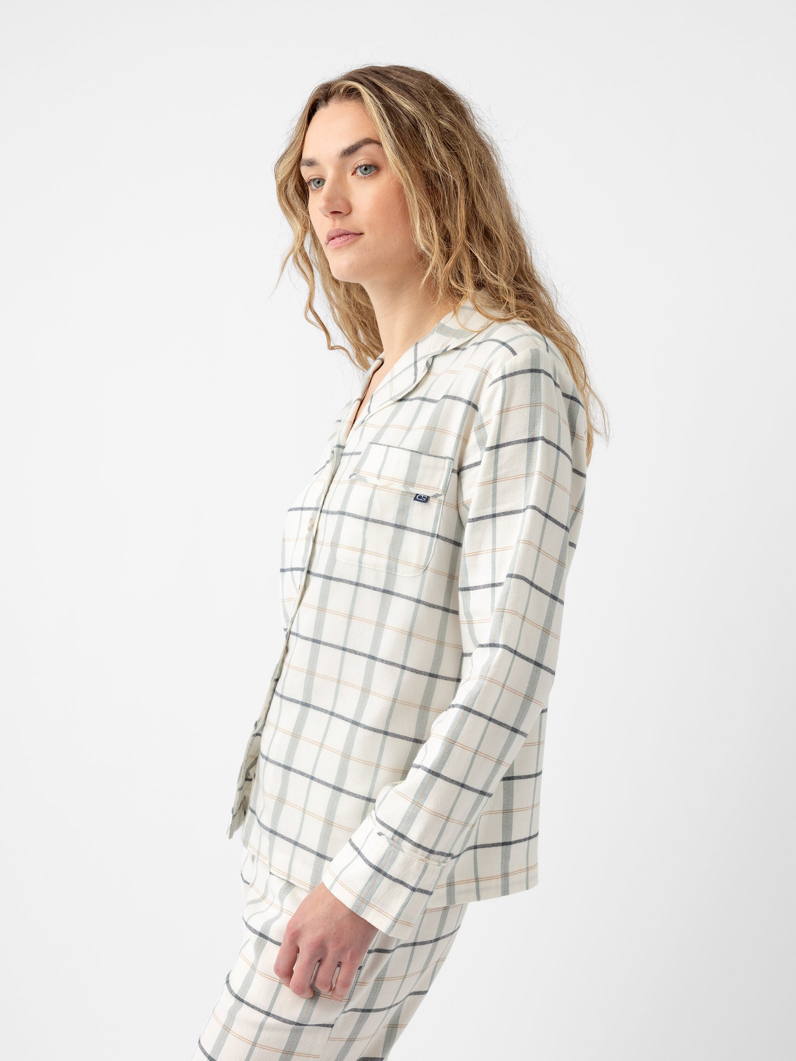 A person with long, wavy hair is wearing the Women's Farmhouse Long Sleeve Pajama Top by Cozy Earth. They are looking to the side, standing against a plain white background. The pajama top features a white and light blue plaid design, long sleeves, and a chest pocket. 